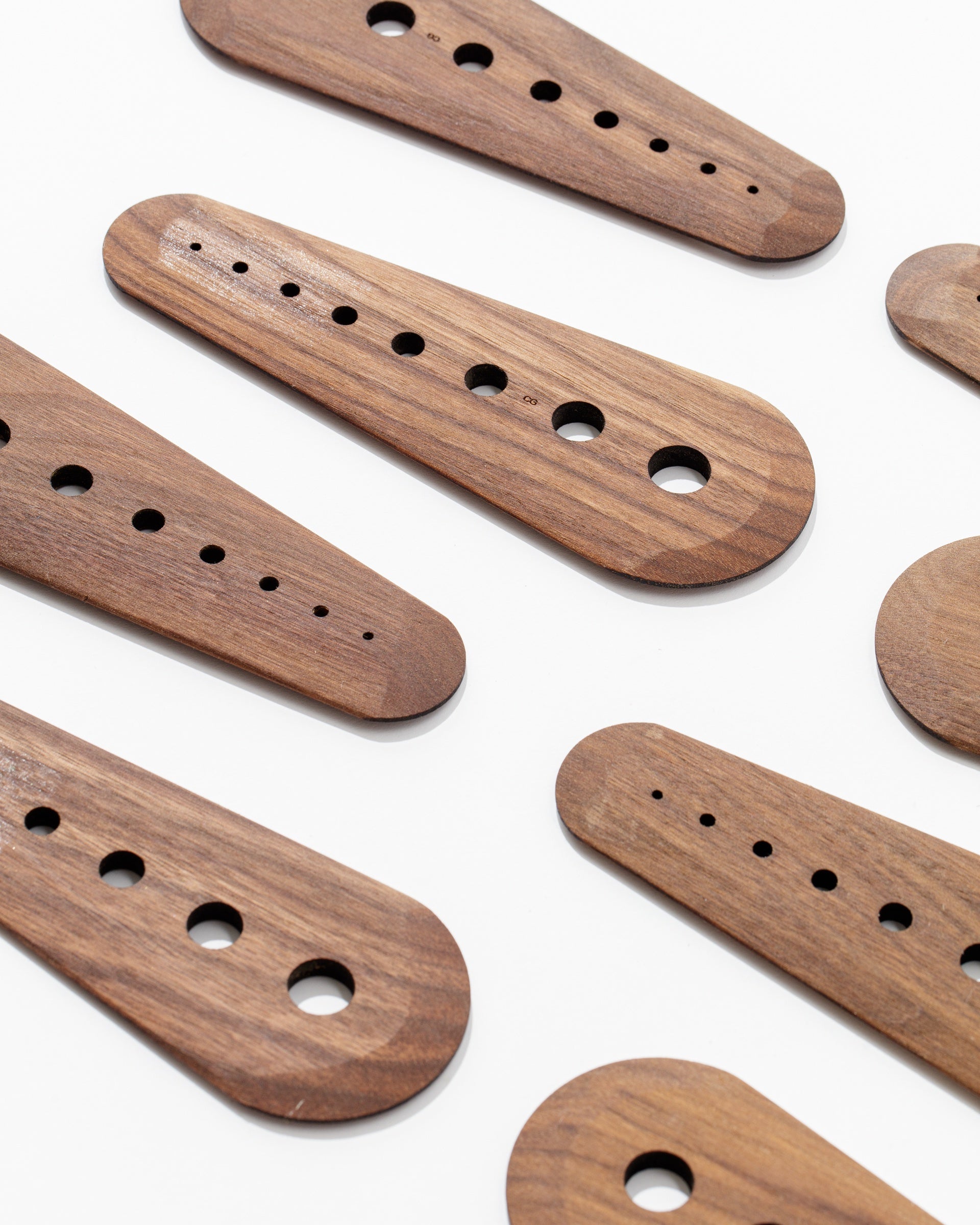 A scattered arrangement of handcrafted Faire Herb Strippers with rounded edges, featuring rows of differently sized circular holes. Each herb stripper is uniformly crafted from dark-stained black walnut wood and displayed against a plain white background.