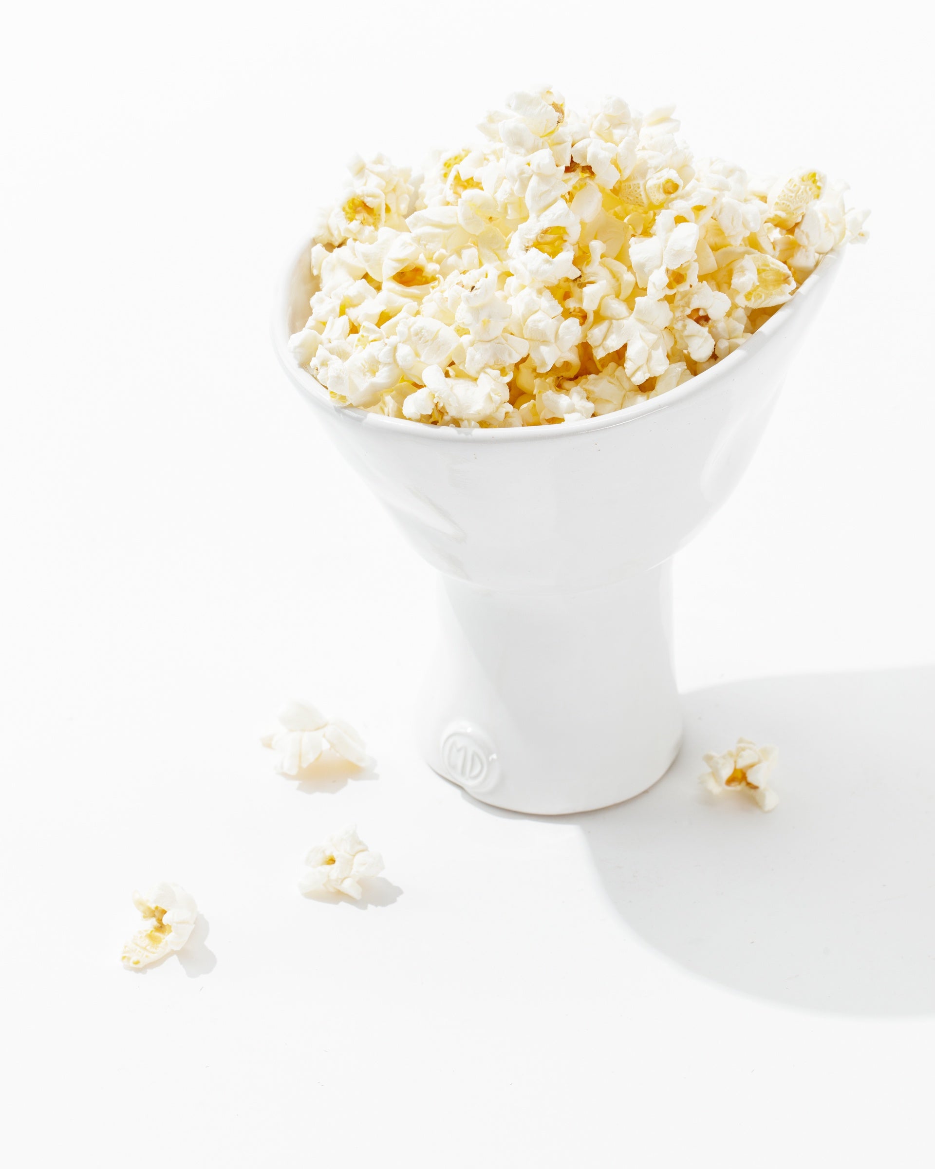 A hand-formed Montes Doggett 5147 catchall bowl no. 147 filled with freshly popped popcorn sits on a clean white surface. Several popped kernels are scattered around the base of the high-fired ceramic bowl, adding a casual, inviting touch to the minimalist composition. Shadows are cast slightly to the right.