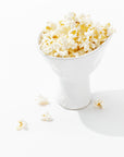 A hand-formed Montes Doggett 5147 catchall bowl no. 147 filled with freshly popped popcorn sits on a clean white surface. Several popped kernels are scattered around the base of the high-fired ceramic bowl, adding a casual, inviting touch to the minimalist composition. Shadows are cast slightly to the right.