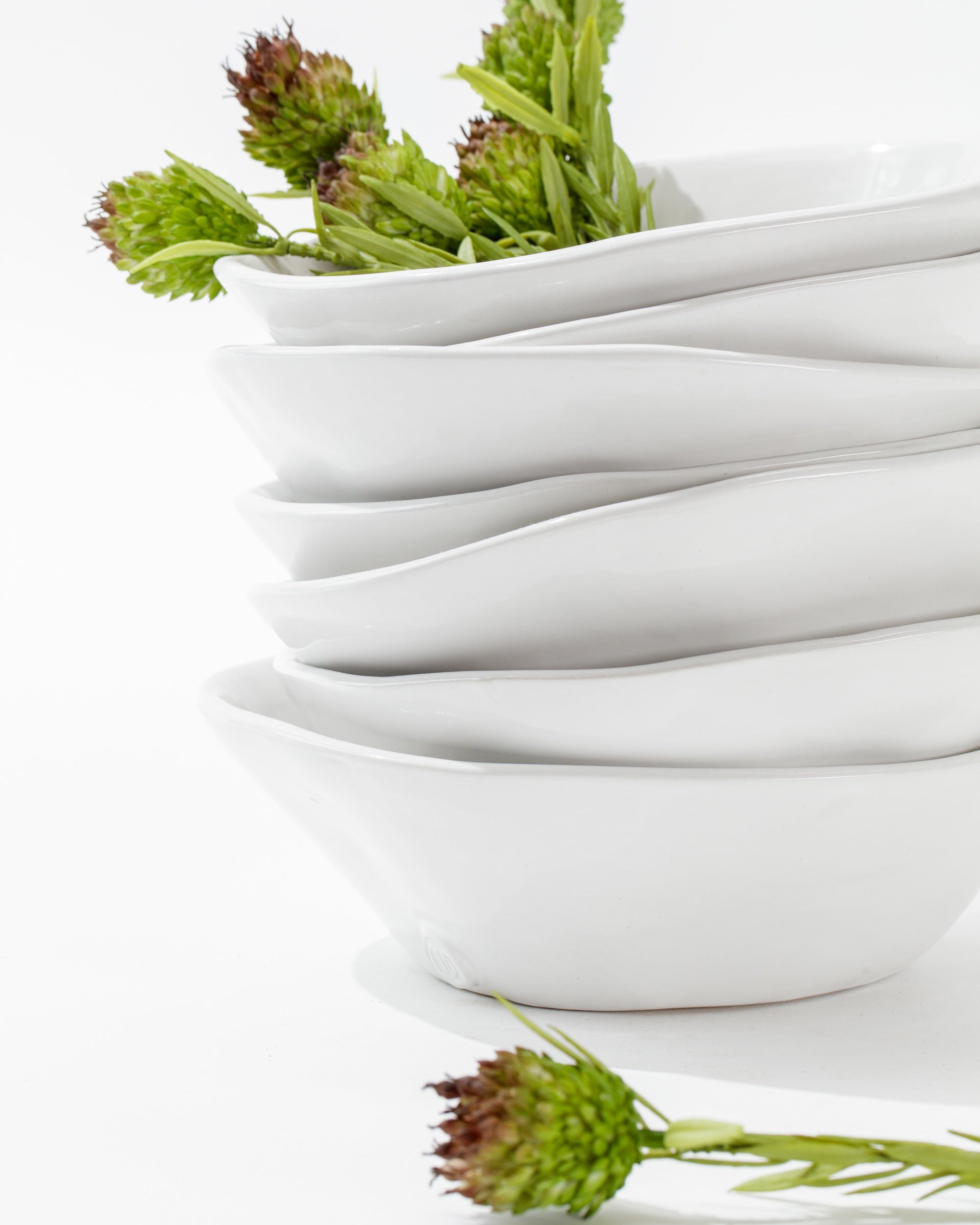 A stack of Montes Doggett&#39;s 5204-L Bowl No. 204 L, featuring white, modern, asymmetrical handmade ceramic design, with a few green spiky flower buds placed inside and leaning against the bowls on a white background. The bowls are arranged in an aesthetically pleasing and curvy manner.