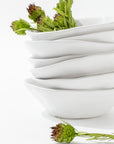 A stack of Montes Doggett's 5204-L Bowl No. 204 L, featuring white, modern, asymmetrical handmade ceramic design, with a few green spiky flower buds placed inside and leaning against the bowls on a white background. The bowls are arranged in an aesthetically pleasing and curvy manner.