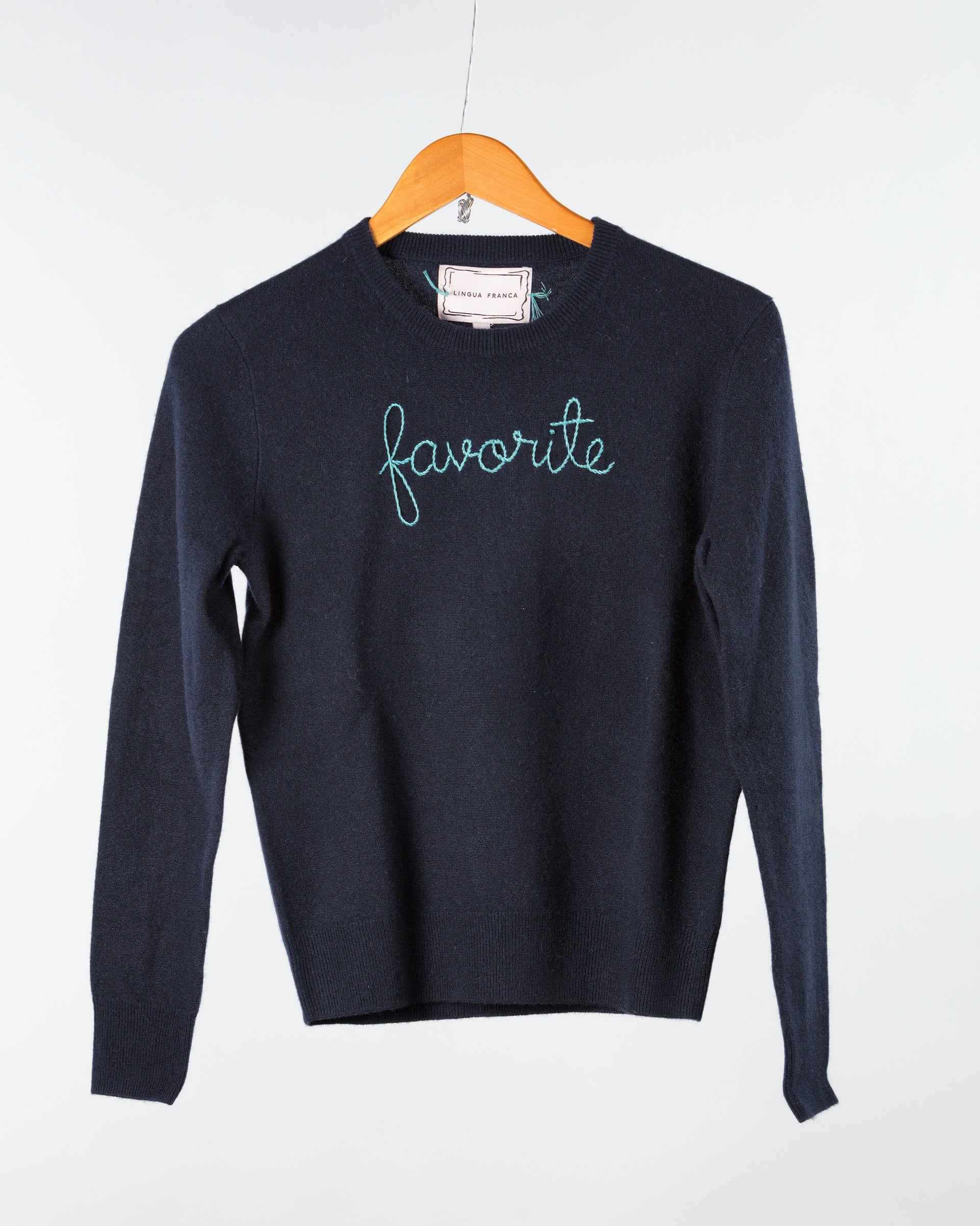 A Lingua Franca navy Cashmere Crewneck sweats on a wooden hanger against a light backdrop, embroidered with "favorite" in light blue script across the chest, underscoring its sustainable origins.