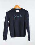 A Lingua Franca navy Cashmere Crewneck sweats on a wooden hanger against a light backdrop, embroidered with "favorite" in light blue script across the chest, underscoring its sustainable origins.