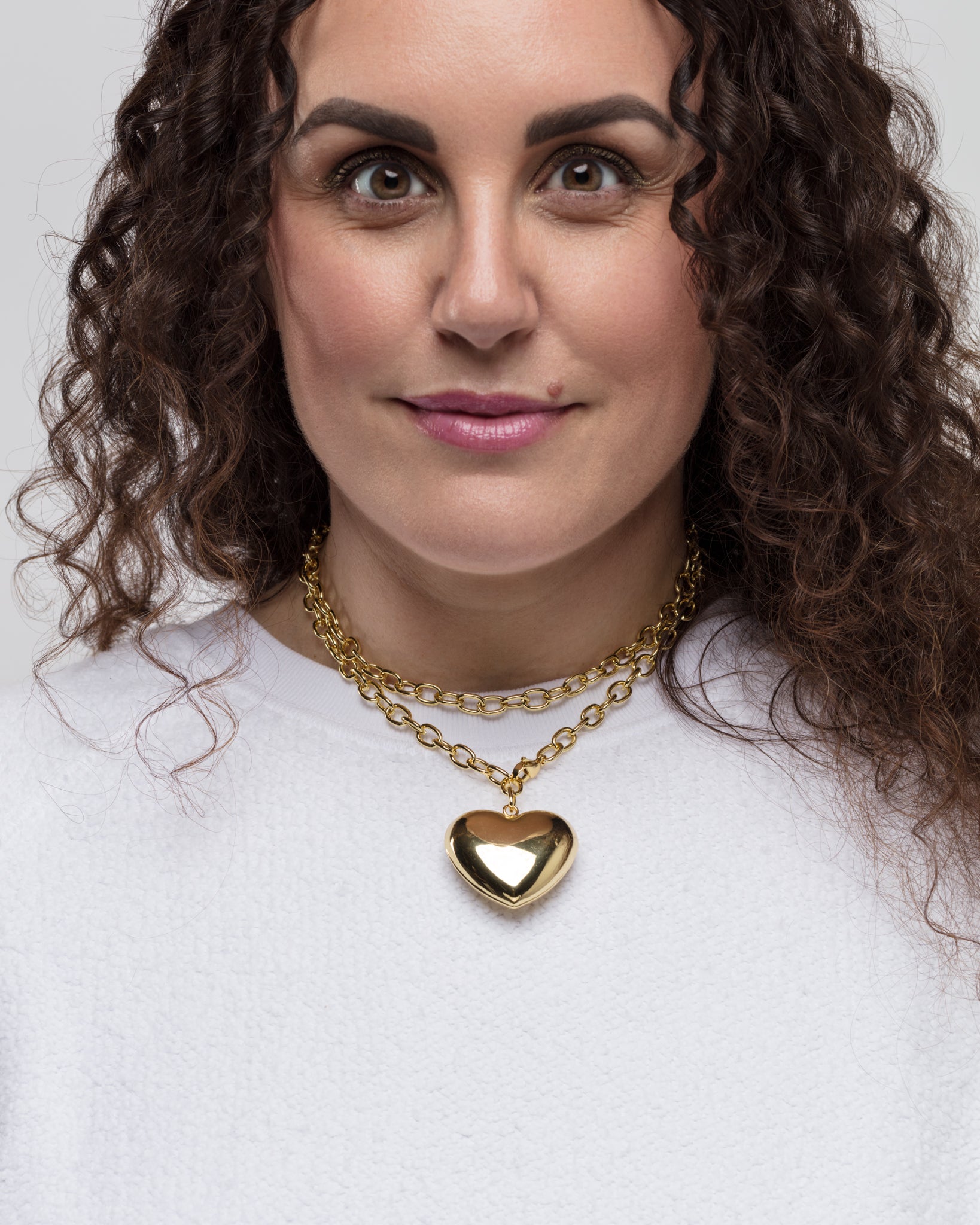 A woman with long, curly dark hair wearing a white top and a Lucky Star Bubble Heart Necklace. She looks directly at the camera with a slight smile.