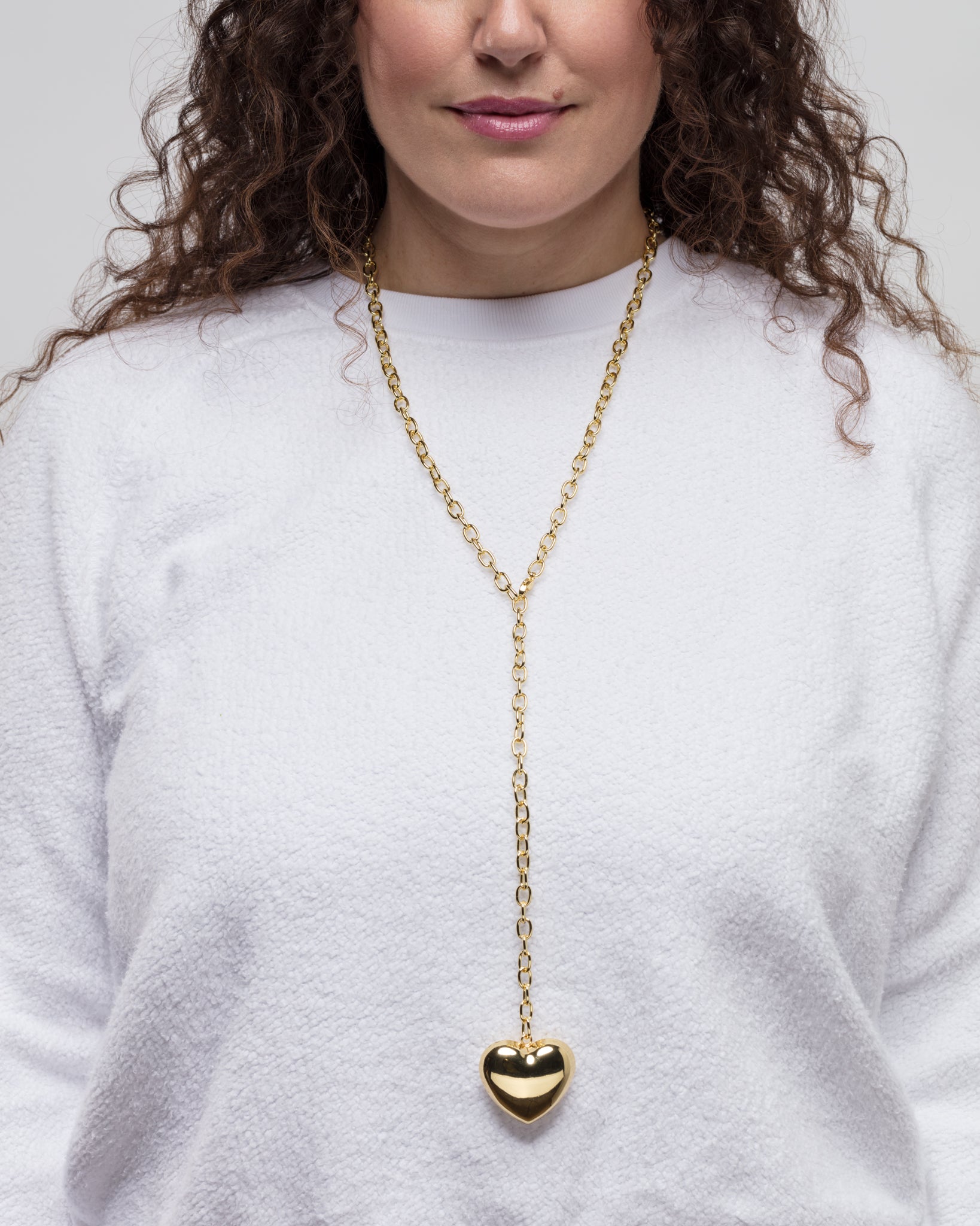 A woman with curly brown hair wearing a white long-sleeved top is shown from the shoulders up. She has a gold chain necklace with the Lucky Star Bubble Heart Necklace, heart-shaped, dangling in front. Her lips are painted a soft pink. The background is plain and light-colored.