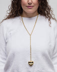 A woman with curly brown hair wearing a white long-sleeved top is shown from the shoulders up. She has a gold chain necklace with the Lucky Star Bubble Heart Necklace, heart-shaped, dangling in front. Her lips are painted a soft pink. The background is plain and light-colored.