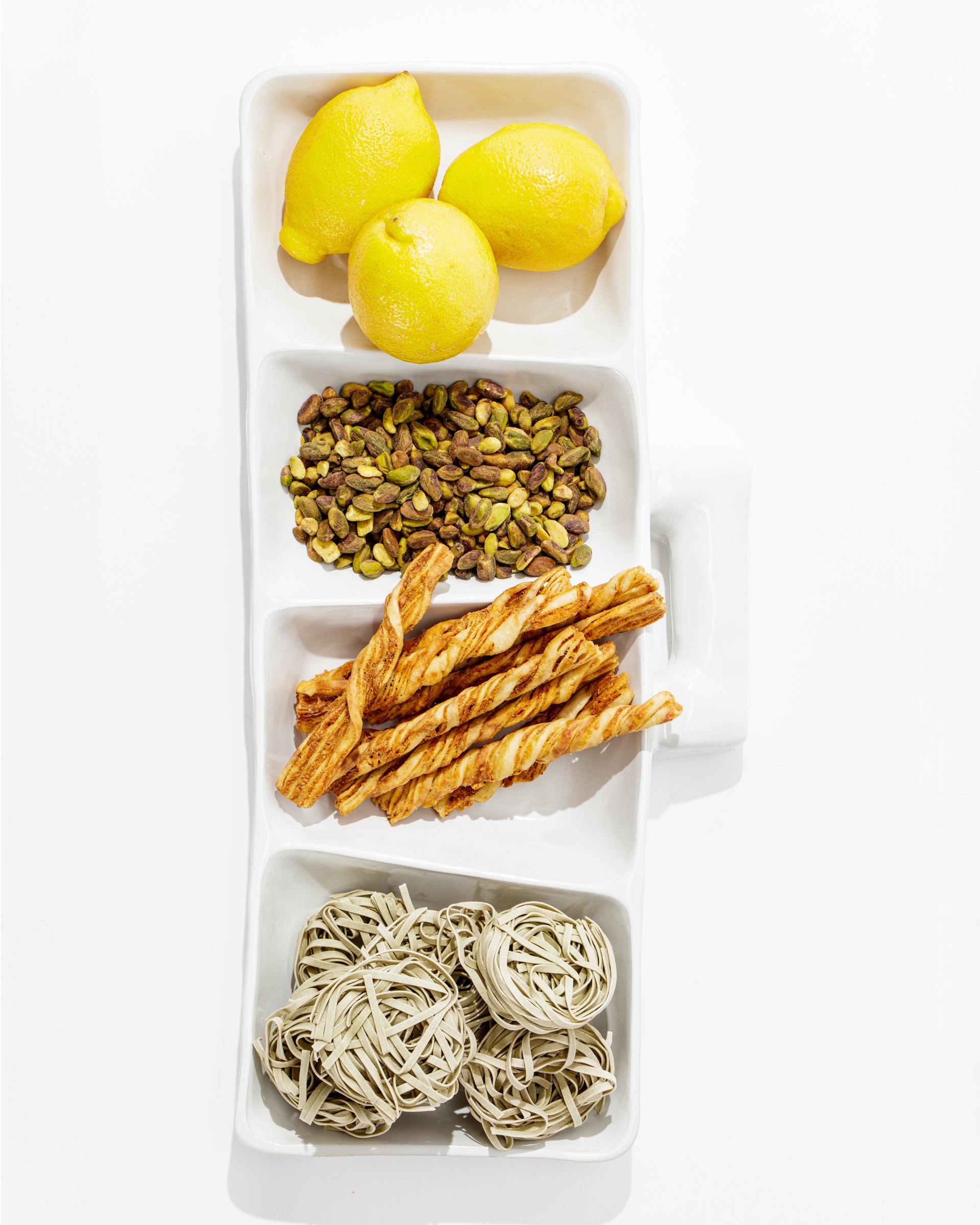 A rectangular white Montes Doggett 5455 Appetizer Platform No. 455 contains yellow lemons in the top section, shelled pistachios beneath them, twisted handmade breadsticks in the third section, and nests of dried pasta in the bottom section.