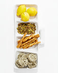 A rectangular white Montes Doggett 5455 Appetizer Platform No. 455 contains yellow lemons in the top section, shelled pistachios beneath them, twisted handmade breadsticks in the third section, and nests of dried pasta in the bottom section.