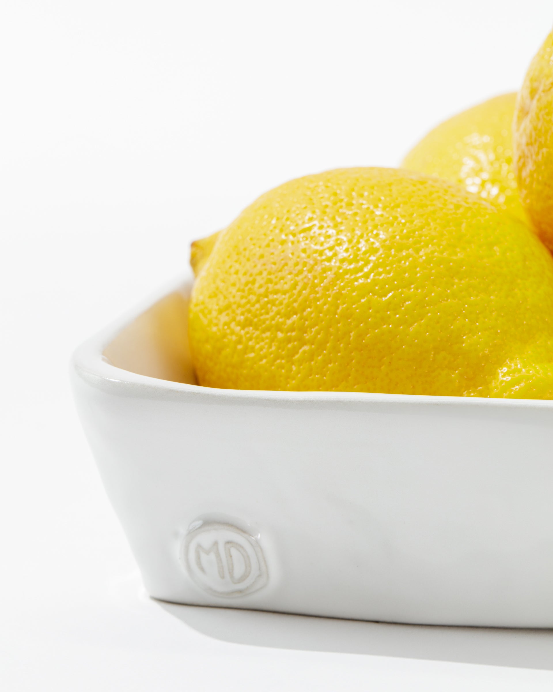 A close-up of a few bright yellow lemons partially visible in the Montes Doggett 5455 Appetizer Platform No. 455, a handmade white ceramic dish with the initials &quot;MD&quot; embossed on its side. The background is white and minimalistic.