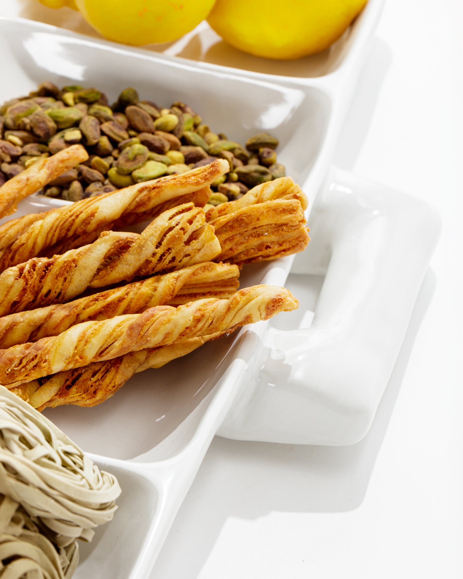 Close-up of the Montes Doggett 5455 Appetizer Platform No. 455, a handmade white ceramic dish with compartments containing various foods. One section contains lemon-yellow fruits, another holds a mixture of pistachio nuts, and the third section showcases twisted breadsticks. The corner of a beige object is partially visible in the foreground.