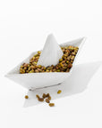 A white, origami-inspired porcelain bowl from Montes Doggett's 5419 Sail Boat no. 419, shaped like a boat and filled with shelled pistachios. Some pistachios are scattered outside the bowl on a white surface. This high-fired ceramic piece, hand made in Peru, artistically mimics the look of a paper boat.