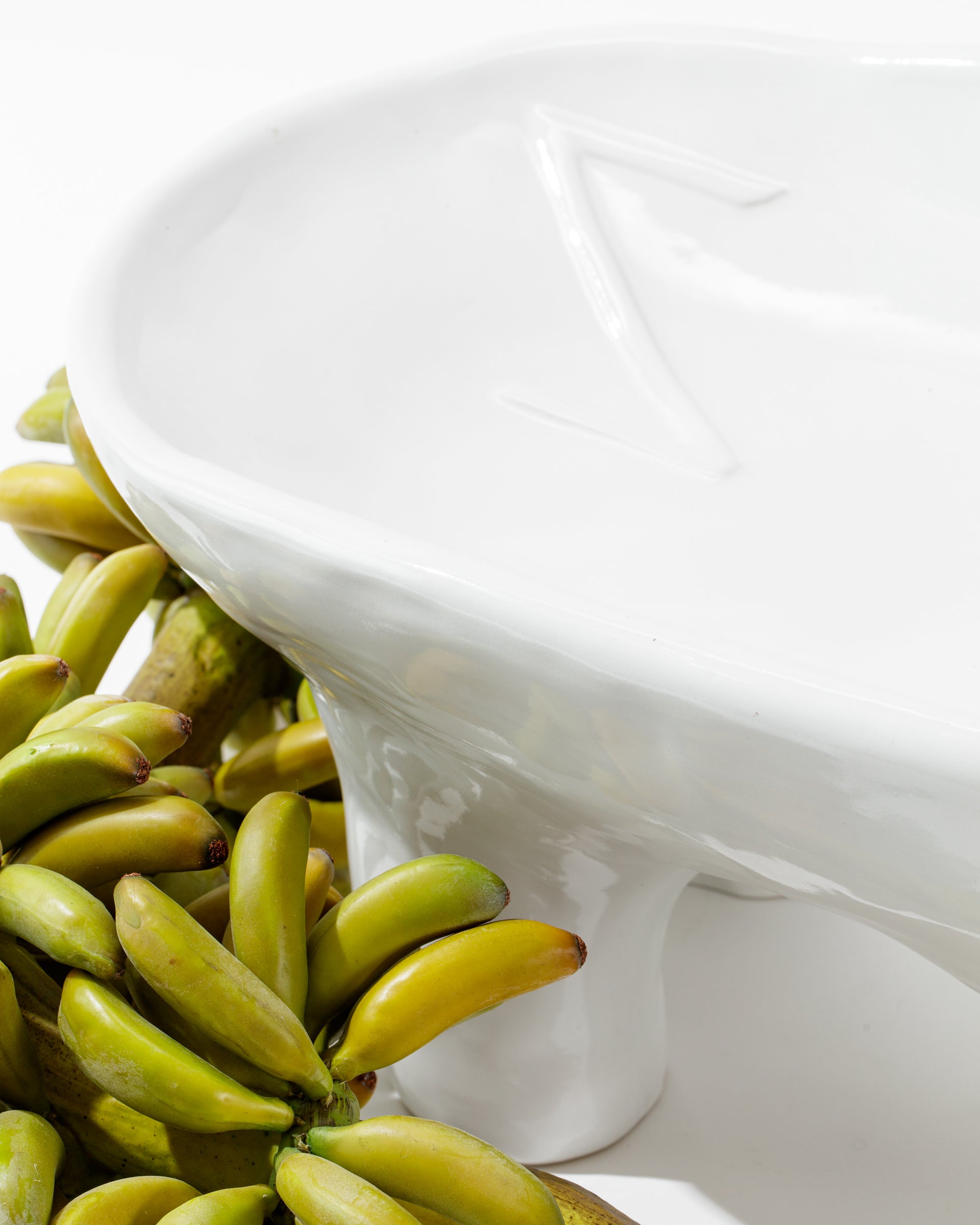 Positioned next to a bunch of vibrant green bananas, the glossy white ceramic 4846 Bowl No.846 by Montes Doggett, featuring an embossed &#39;Z&#39; and handmade in Peru, showcases a modern, minimalist design. The bowl stands on two small legs while the bananas are artfully arranged to its left.