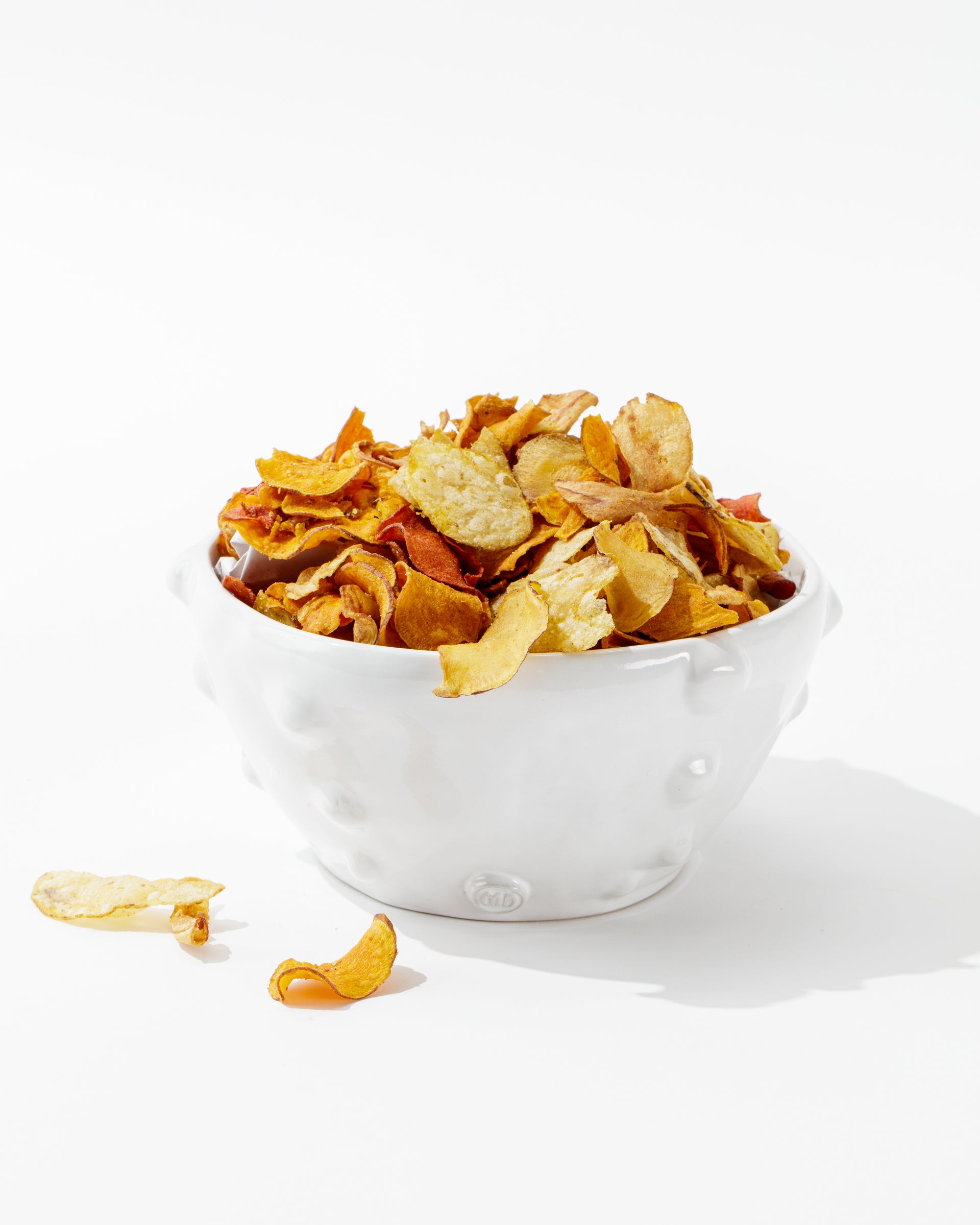 Bowl No. 130, a handmade, white, textured ceramic creation by Montes Doggett, is filled with mixed vegetable chips that showcase a variety of colors and textures. A few chips are scattered on the white surface beside the bowl. The background is clean and bright, emphasizing the vibrant hues of the chips.