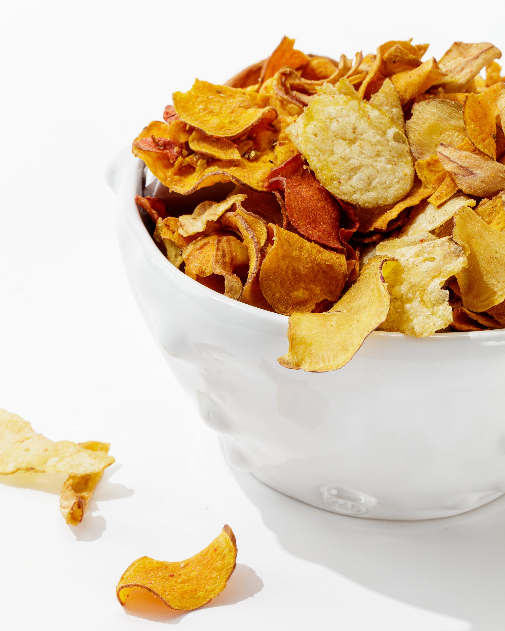 A white handmade Montes Doggett Bowl No. 130 filled with various dried vegetable chips, including sweet potatoes, beets, and parsnips. Some chips are spilled out of the durable high-fired bowl onto a white surface, adding a rustic touch. The vivid hues of orange, red, and yellow stand out against the bowl&#39;s neutral tone.