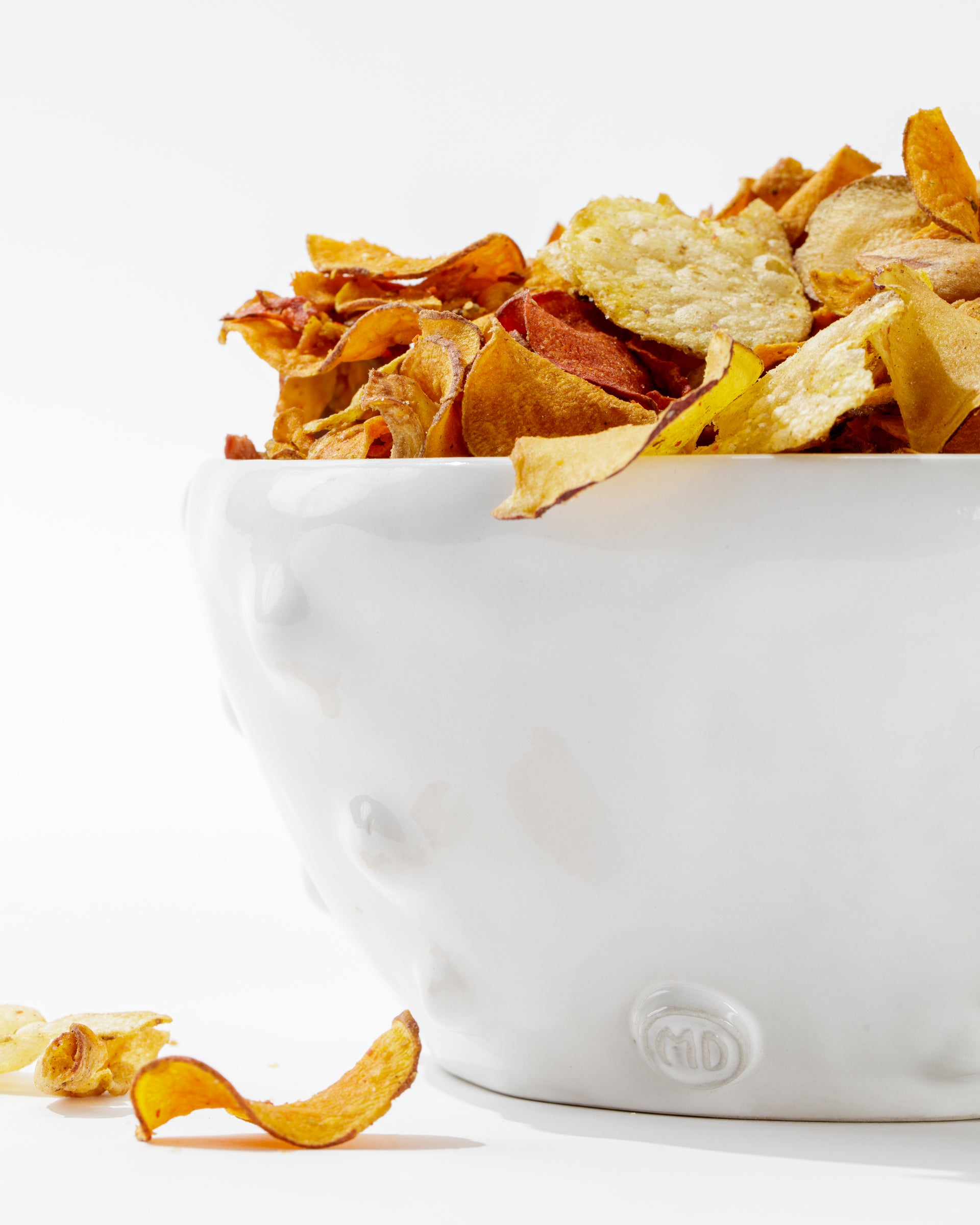 A white, handmade ceramic Bowl No. 130 by Montes Doggett filled with a variety of crispy, golden potato chips. Some chips spill out onto the white surface below, creating a casual and inviting snack scene that showcases traditional techniques.