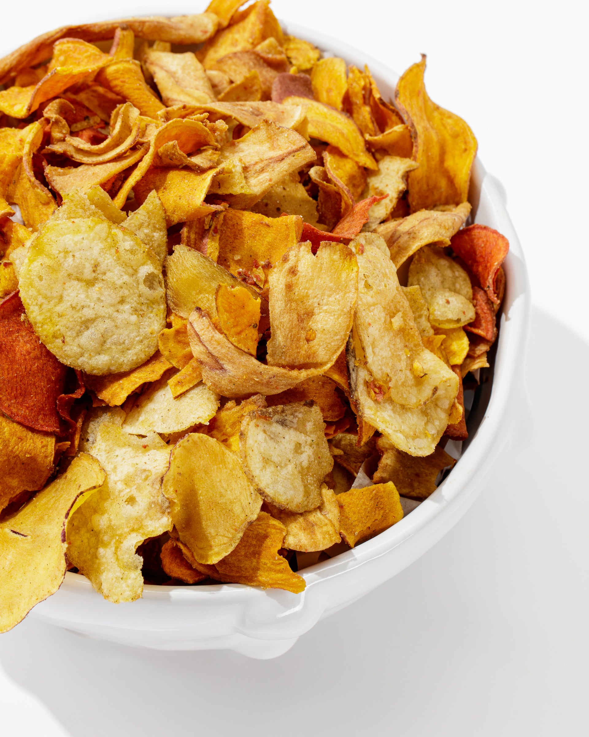 A white handmade Bowl No. 130 by Montes Doggett is filled with a variety of vegetable chips, including sweet potato, beet, and parsnip chips, all against a white background. The chips are golden, orange, and red in color, beautifully showcasing their crispy texture. This durable high-fired piece is crafted using traditional techniques.