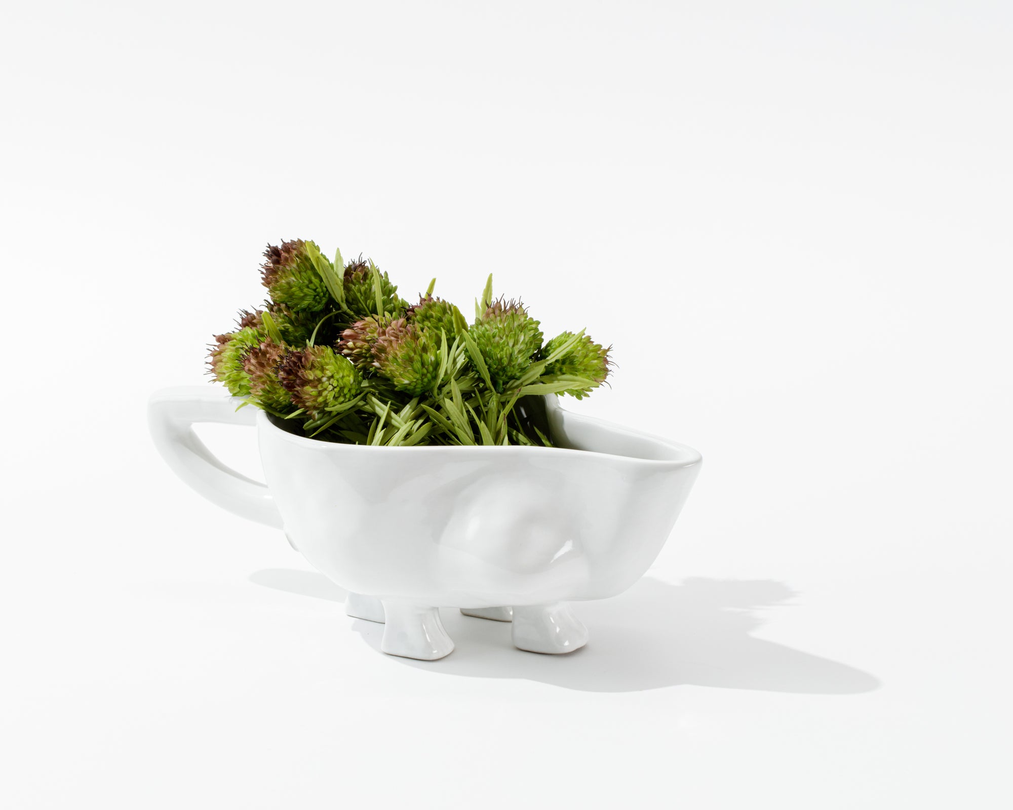 The Montes Doggett Gravy Boat No. 151, a whimsical, white high-fired ceramic container with short legs and a handle, is filled with green chive blossoms. Handmade in Peru, the container casts a soft shadow on the clean, white background.