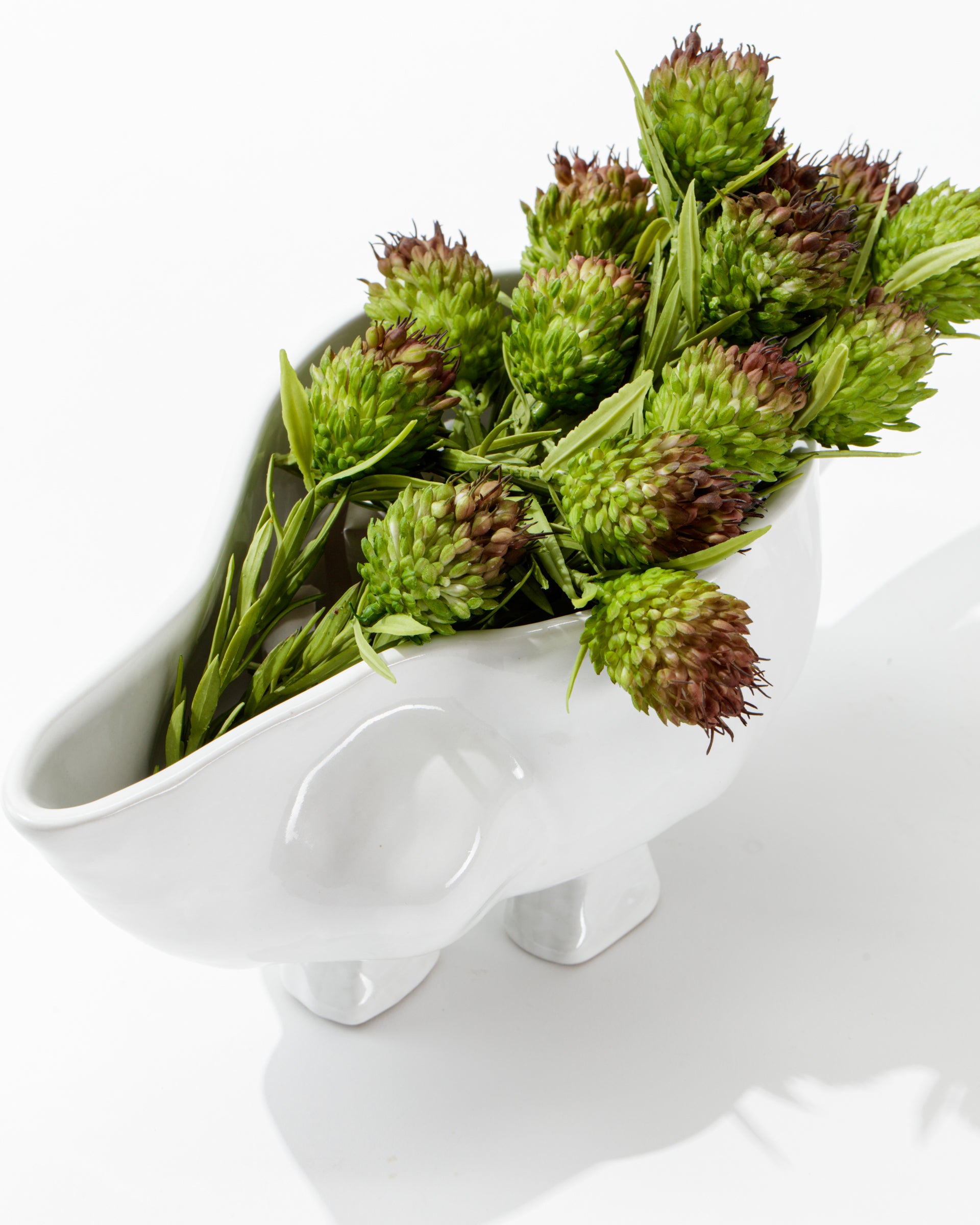 The Montes Doggett Gravy Boat No. 151, a handmade white high-fired ceramic vessel, shaped like an abstract hippo and featuring a glossy finish, contains a bunch of green, spiky flowers with reddish tips. Made in Peru, it stands on four short legs against a clean, bright white background.