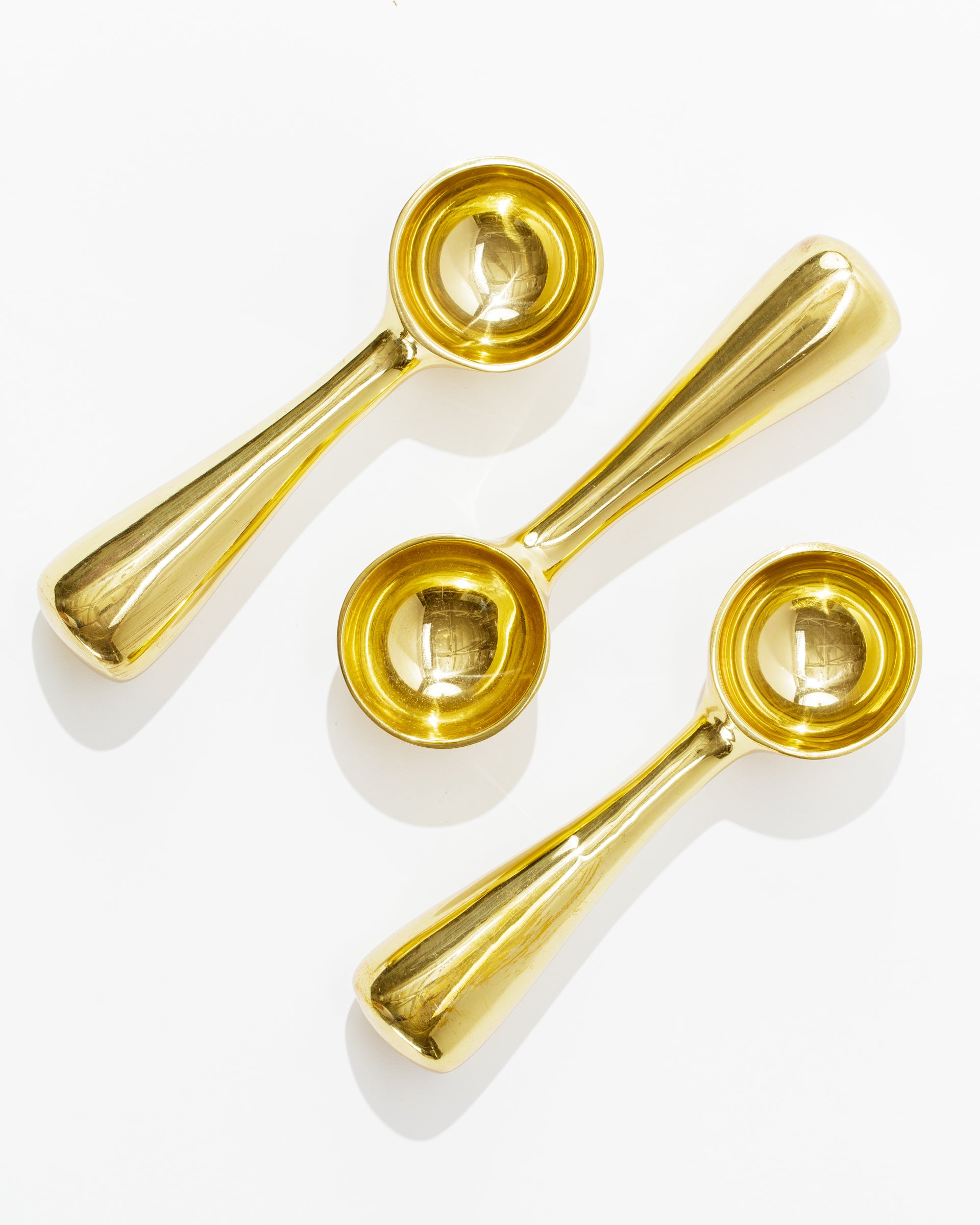 Three shiny, gold-colored Brass Dessert Scoops of varying sizes from Sir/Madam are arranged on a white background. Crafted from 100% food-safe brass, these scoops feature a sleek, modern design with short, thick handles and rounded bowls. Their reflective surfaces create bright highlights, making them an elegant addition to any kitchenalia collection.