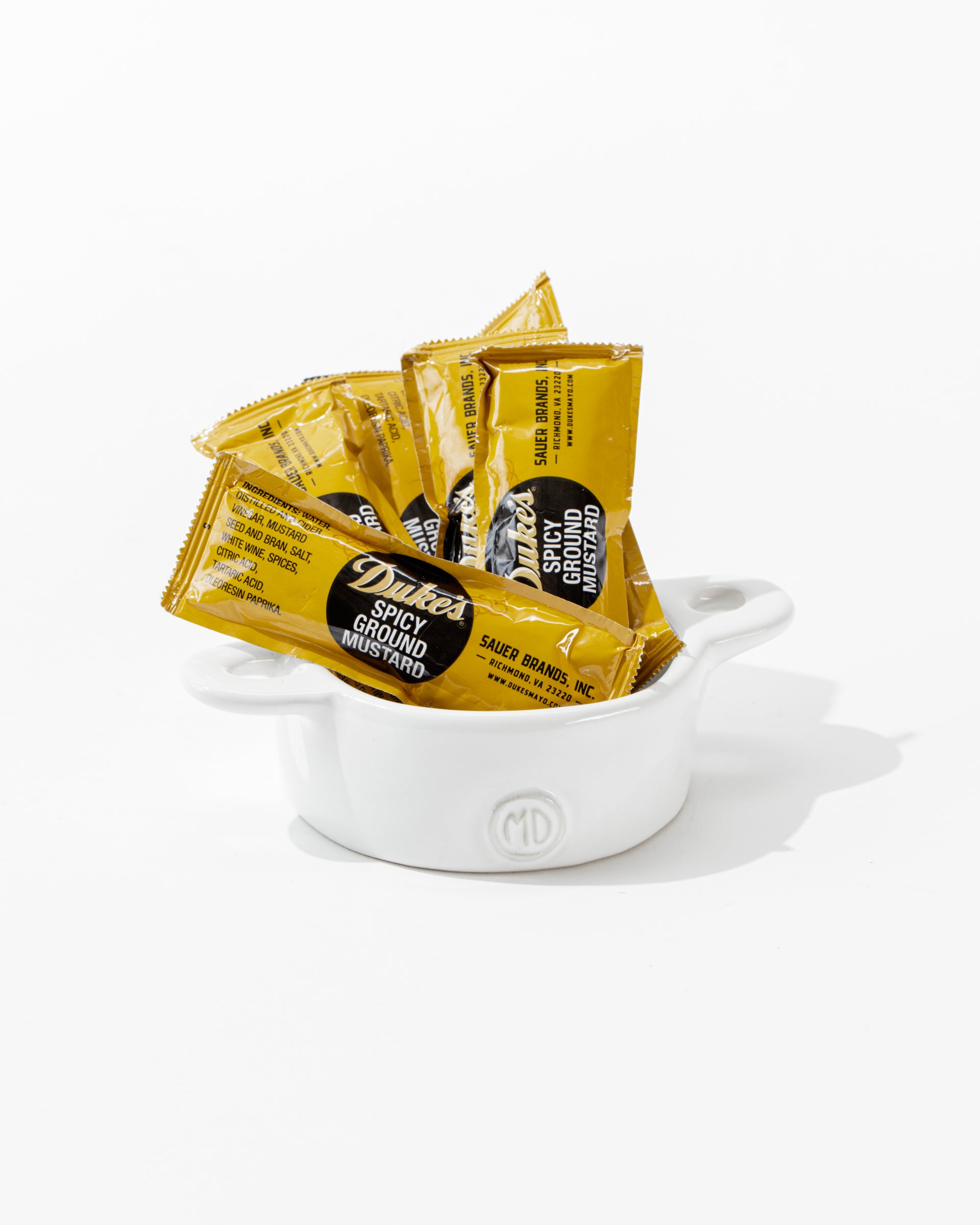 A small white high fired ceramic dish called Bowl No. 5 by Montes Doggett, handmade in Peru, filled with individually wrapped packets of Duke's Spicy Ground Mustard. The yellow packets with black and white labeling are arranged upright in the dish against a plain white background.