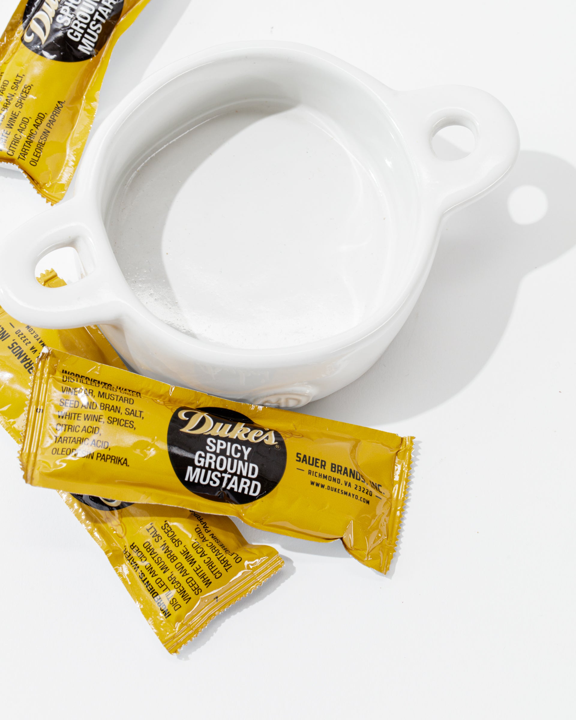 A high-fired ceramic dish, named Bowl No. 5 from Montes Doggett, sits on a white surface with three yellow packets of Duke's Spicy Ground Mustard placed beside and on top of it. The handmade dish, crafted in Peru, complements the sealed packets clearly labeled with ingredient information and branding.