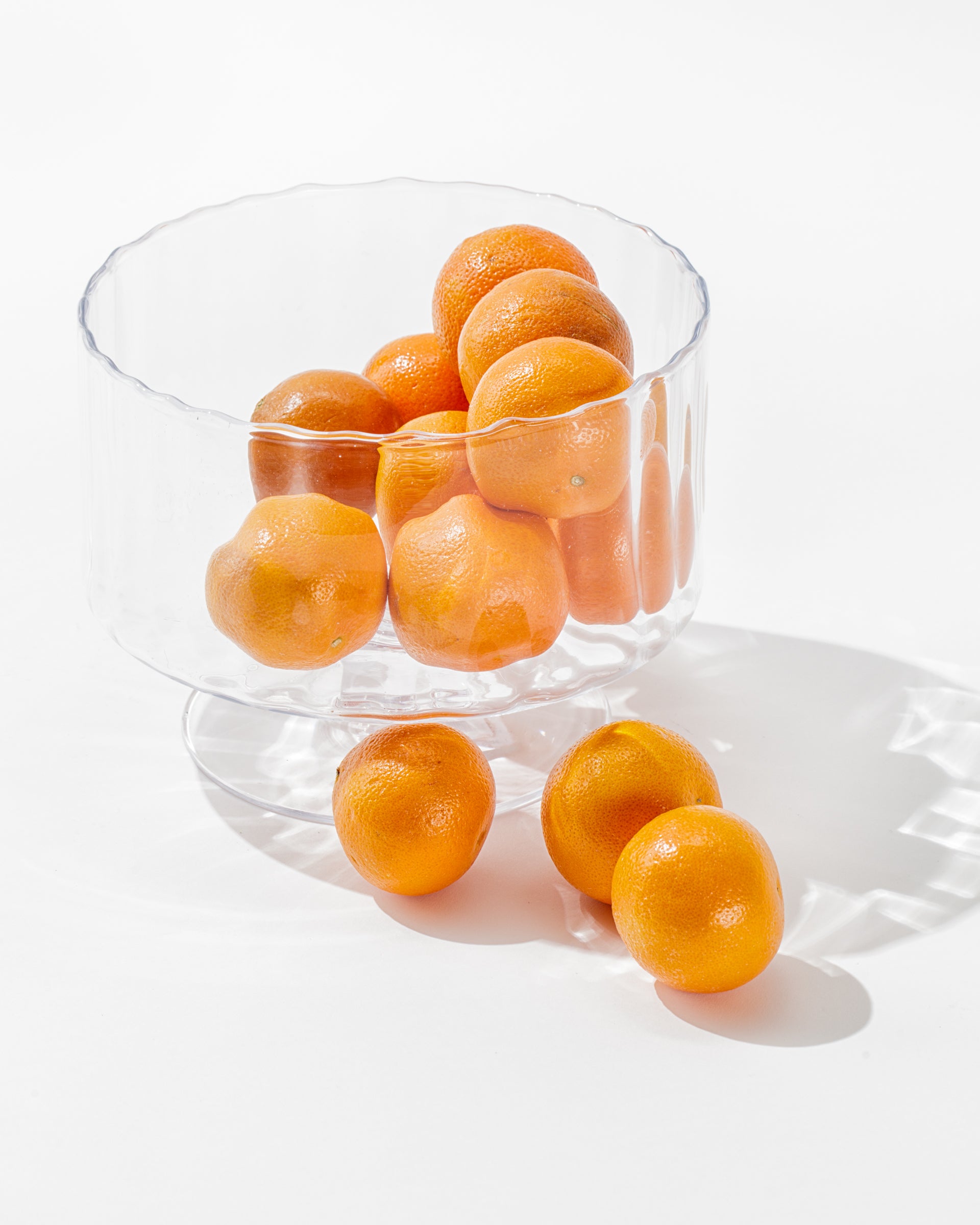 A clear Scalloped Glass Bowl by Zodax, filled with several bright orange clementines, placed on a white surface, with a few clementines scattered outside the bowl. This elegant decor casts gentle shadows, highlighting the smooth texture and vibrant color of the fruit.