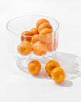 A clear Scalloped Glass Bowl by Zodax, filled with several bright orange clementines, placed on a white surface, with a few clementines scattered outside the bowl. This elegant decor casts gentle shadows, highlighting the smooth texture and vibrant color of the fruit.