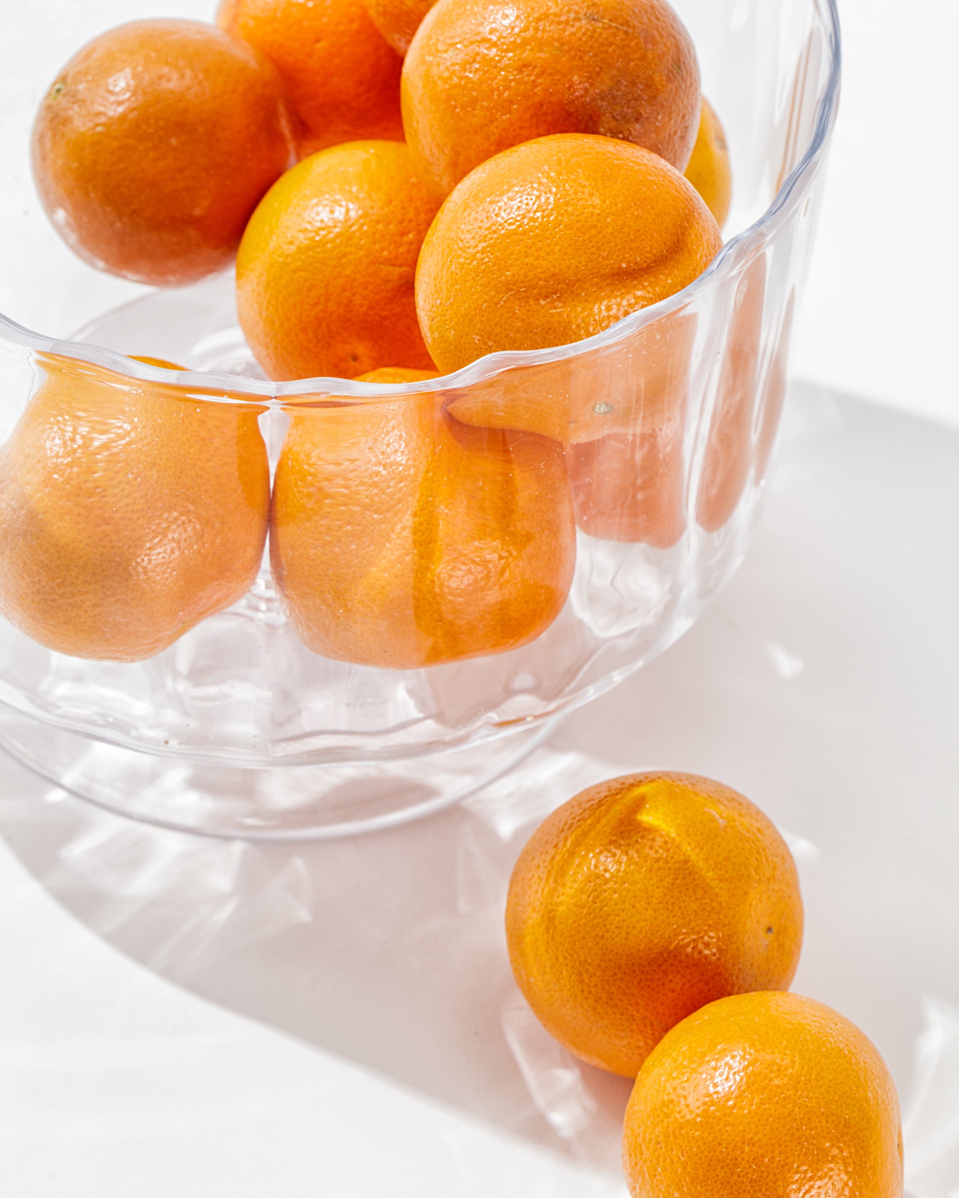 A clear Zodax Scalloped Glass Bowl filled with bright orange tangerines sits on a white surface. Two tangerines are placed outside the bowl, creating a simple and fresh composition. The handcrafted design of the Scalloped Glass Bowl adds to the elegant decor, showcasing the vibrant color and smooth texture of the fruit.