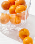 A clear Zodax Scalloped Glass Bowl filled with bright orange tangerines sits on a white surface. Two tangerines are placed outside the bowl, creating a simple and fresh composition. The handcrafted design of the Scalloped Glass Bowl adds to the elegant decor, showcasing the vibrant color and smooth texture of the fruit.