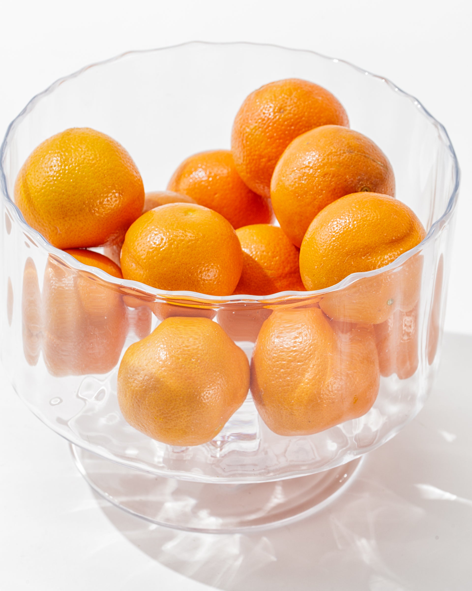 A clear Zodax Scalloped Glass Bowl filled with vibrant, round oranges against a plain white background. The textured scalloped bowl showcases the bright orange color of the fruits, creating an elegant decor and visually appealing composition.