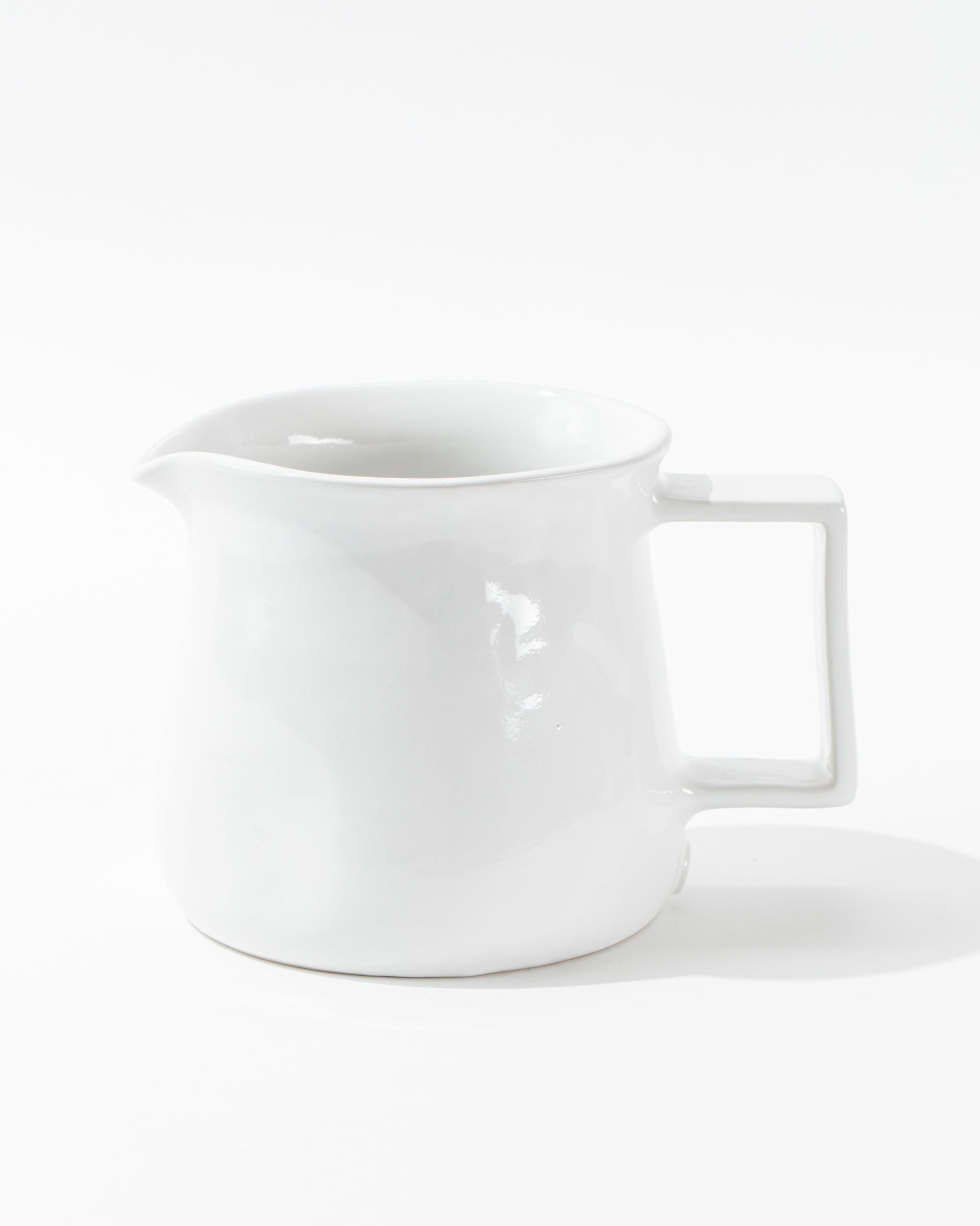 Introducing the Montes Doggett Pitcher No. 750, a high-fired, white ceramic pitcher featuring a square handle and spout. Its design is simple and minimalistic with a glossy finish, and this artisan-crafted piece sits gracefully on a white surface against a plain background.