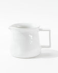 Introducing the Montes Doggett Pitcher No. 750, a high-fired, white ceramic pitcher featuring a square handle and spout. Its design is simple and minimalistic with a glossy finish, and this artisan-crafted piece sits gracefully on a white surface against a plain background.