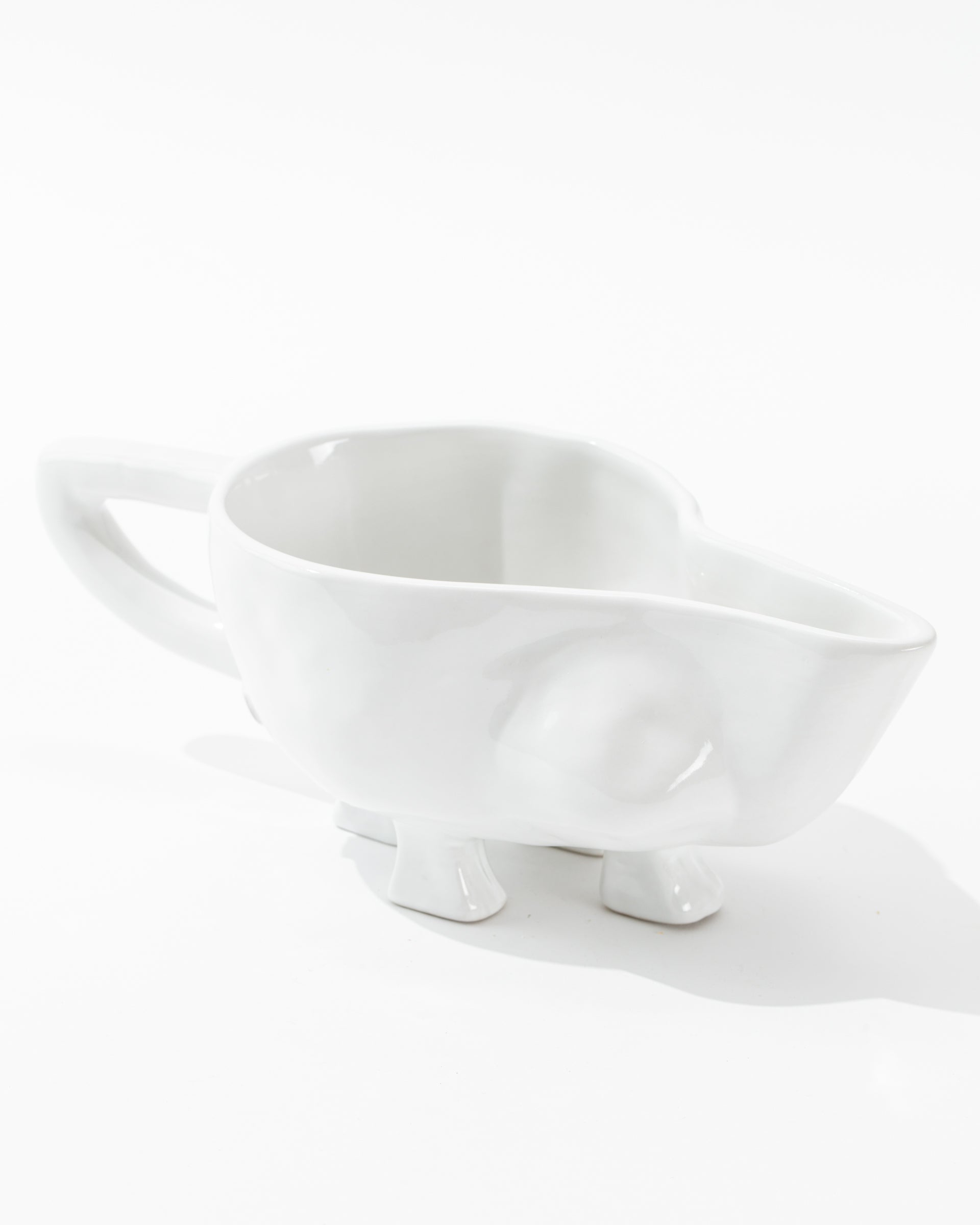 The Gravy Boat No. 151, part of the Montes Doggett ceramic collection, is a handmade white ceramic piece shaped like an elephant, featuring a handle that resembles its trunk. Crafted from high-fired ceramic, it boasts minimalistic details such as two small ears and four stubby legs as its base, giving it a whimsical and artistic design.