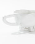 The Gravy Boat No. 151 by Montes Doggett is a quirky ceramic piece shaped like an animal with four leg-like feet and an oversized handle. Its minimalist design, with no distinctive features beyond the feet and handle, showcases the beauty of high-fired ceramic craftsmanship as it rests on a flat white surface.