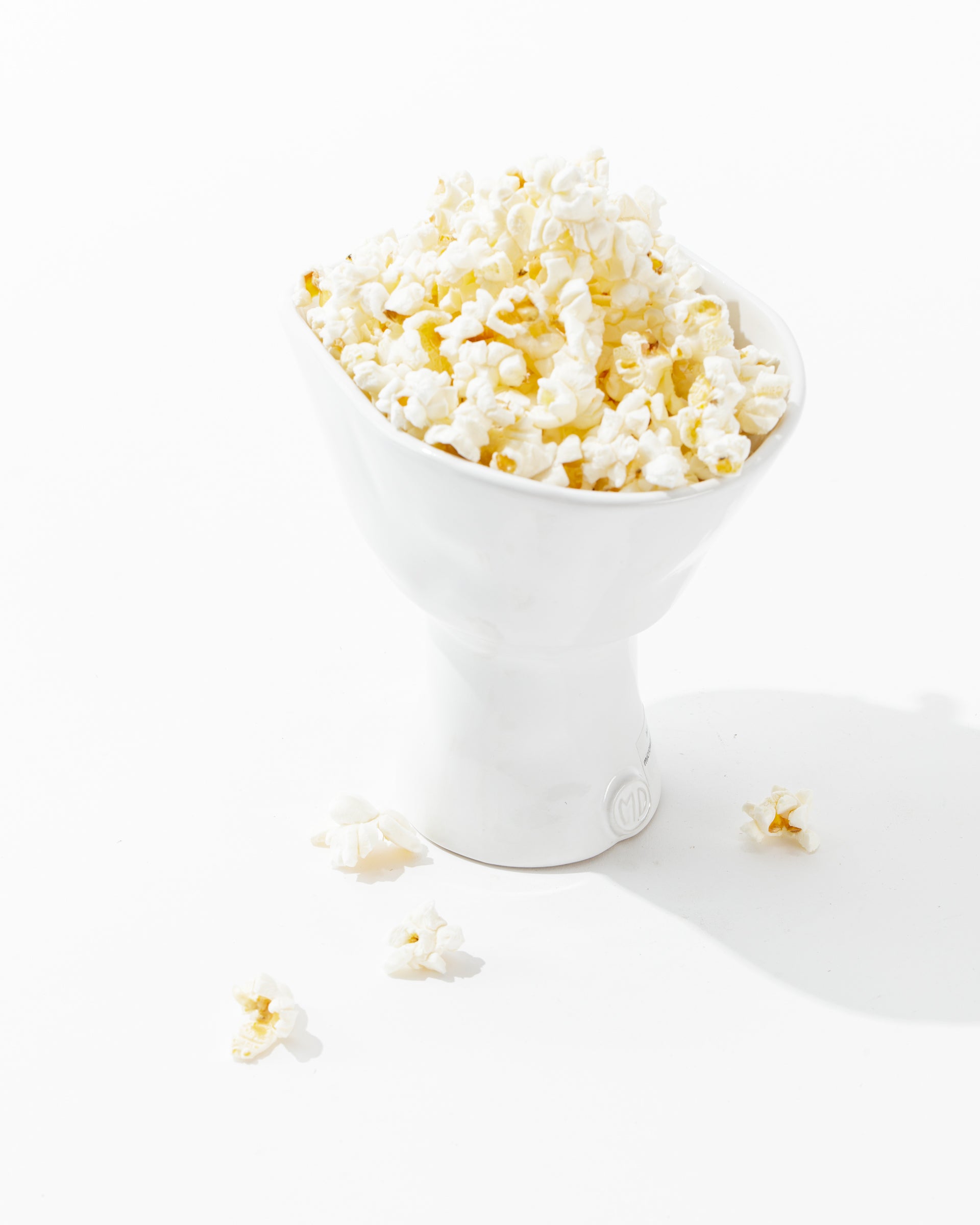 The 5147 catchall bowl no. 147 by Montes Doggett, filled with popped popcorn kernels and a few pieces scattered around the base, stands on a white surface. The image is brightly lit, showcasing the texture and light color of the popcorn.