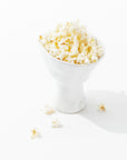 The 5147 catchall bowl no. 147 by Montes Doggett, filled with popped popcorn kernels and a few pieces scattered around the base, stands on a white surface. The image is brightly lit, showcasing the texture and light color of the popcorn.