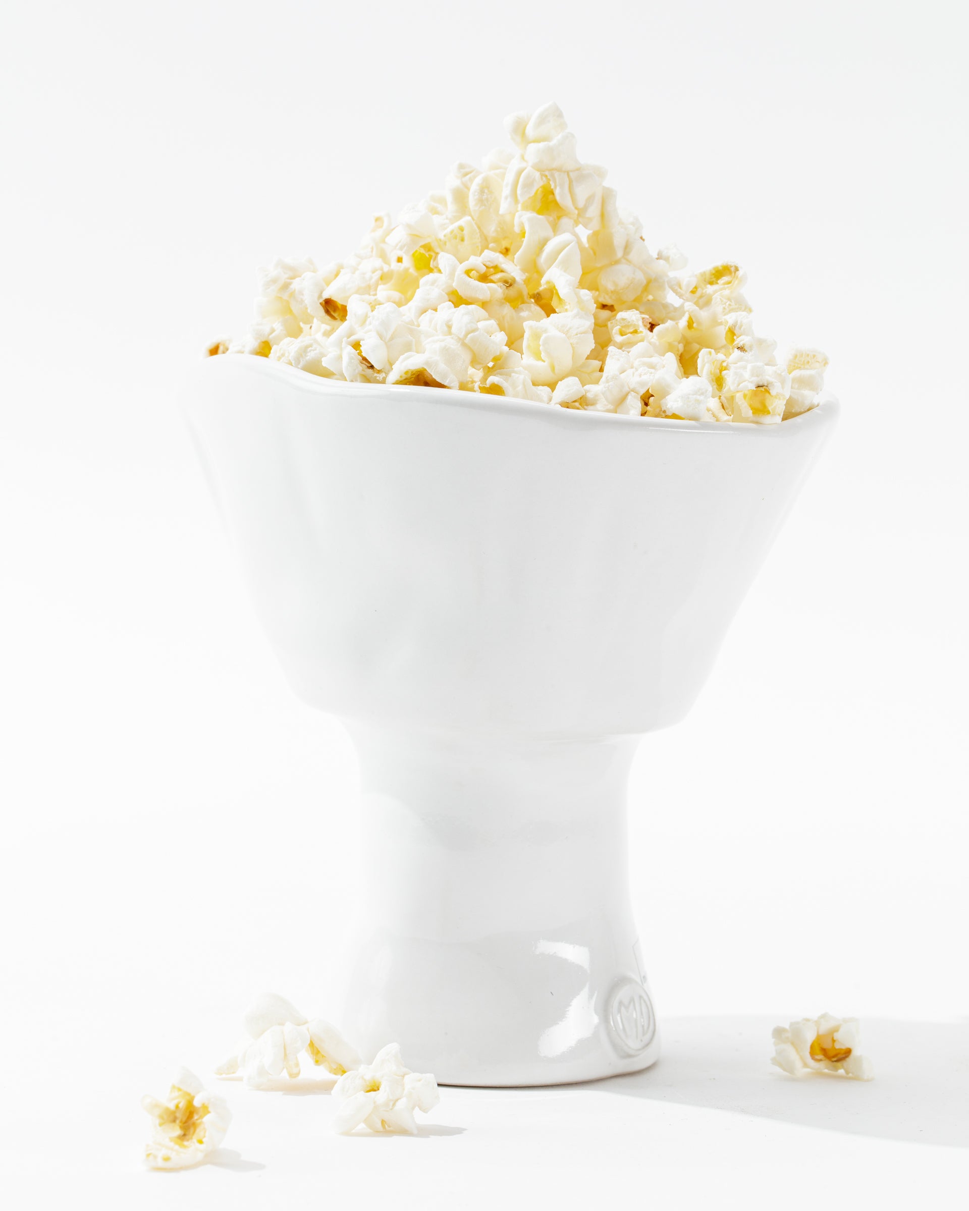 A sleek, white, modern catchall bowl handcrafted by skilled artisans, the 5147 Catchall Bowl No. 147 from Montes Doggett, is filled to the brim with fluffy, freshly popped popcorn. A few kernels have spilled onto the white surface below, creating a clean and minimalist aesthetic. The scene is brightly lit, emphasizing the texture of the popcorn.