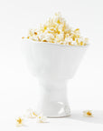 A sleek, white, modern catchall bowl handcrafted by skilled artisans, the 5147 Catchall Bowl No. 147 from Montes Doggett, is filled to the brim with fluffy, freshly popped popcorn. A few kernels have spilled onto the white surface below, creating a clean and minimalist aesthetic. The scene is brightly lit, emphasizing the texture of the popcorn.