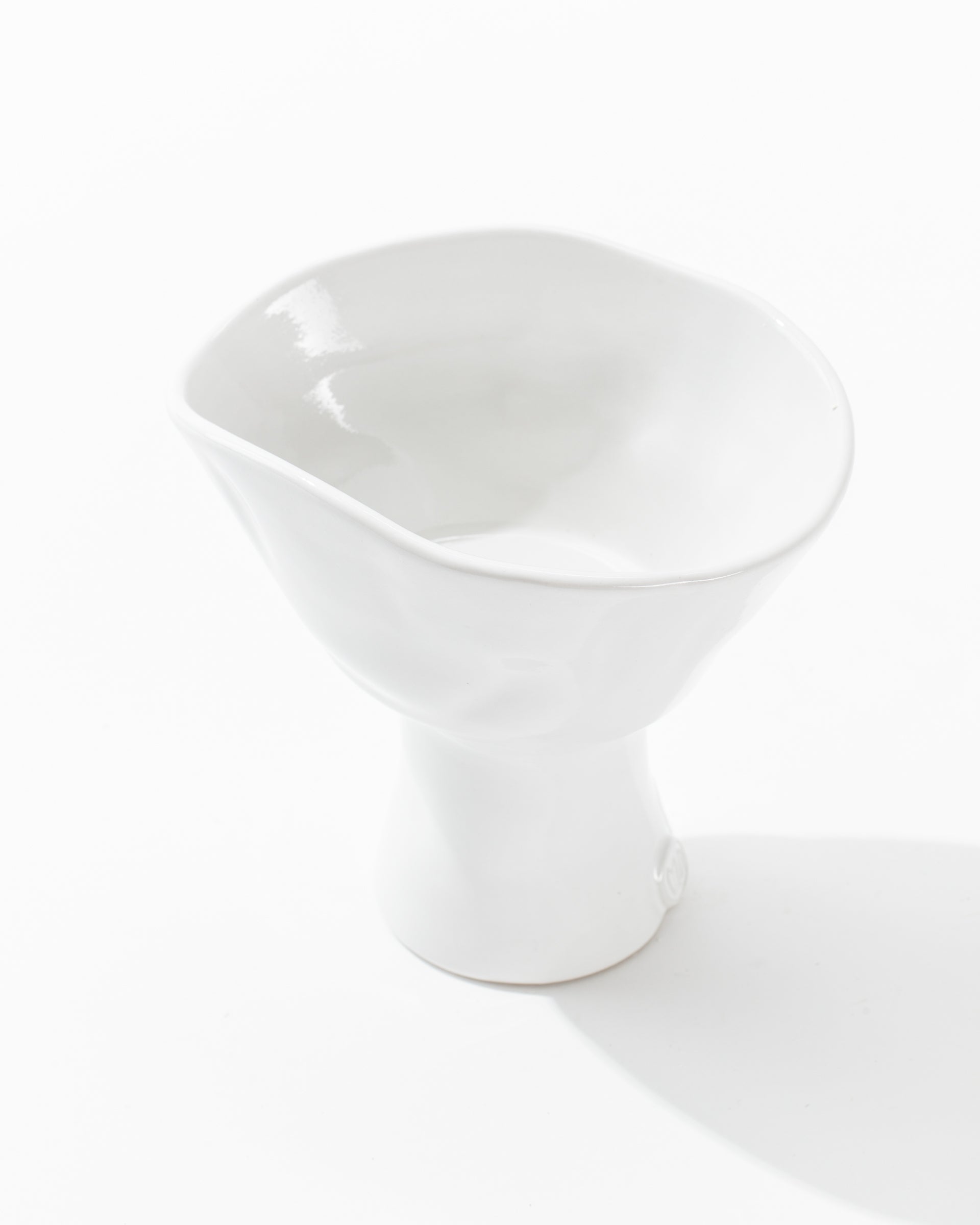 Introducing the 5147 Catchall Bowl No. 147 by Montes Doggett, a sleek and asymmetrical white ceramic piece with a glossy finish. The top features a unique, tilted and irregular rim, while the base is more narrow and cylindrical in shape. Meticulously crafted by skilled artisans, this bowl highlights exceptional durability against its plain white backdrop.