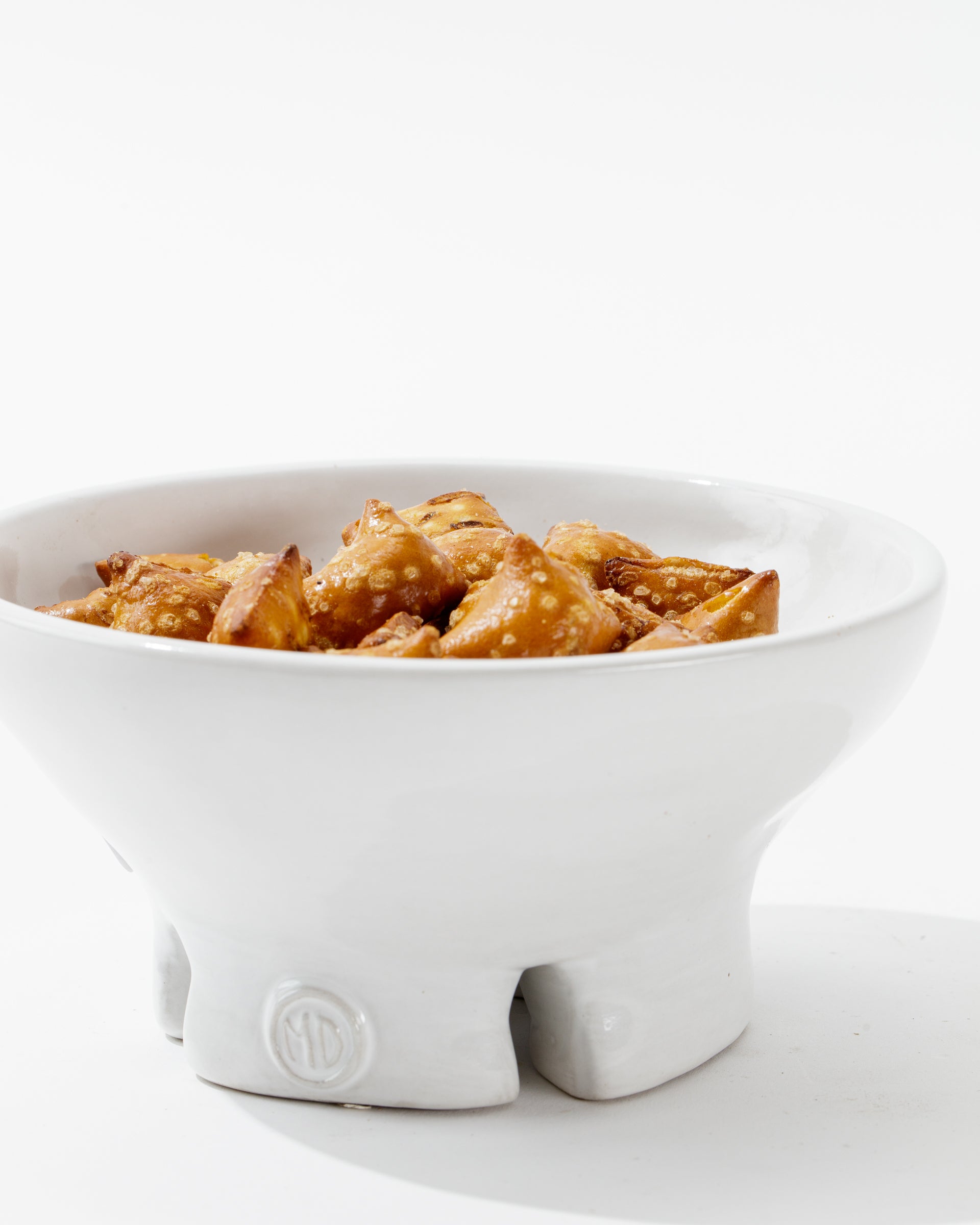 The Catchall Bowl No. 257 by Montes Doggett, crafted using traditional techniques from high fired clay, contains golden-brown fried appetizers, possibly samosas or wontons. Placed against a plain white background, this handmade ceramic dinnerware boasts a unique design with three short legs and a small emblem on the front.