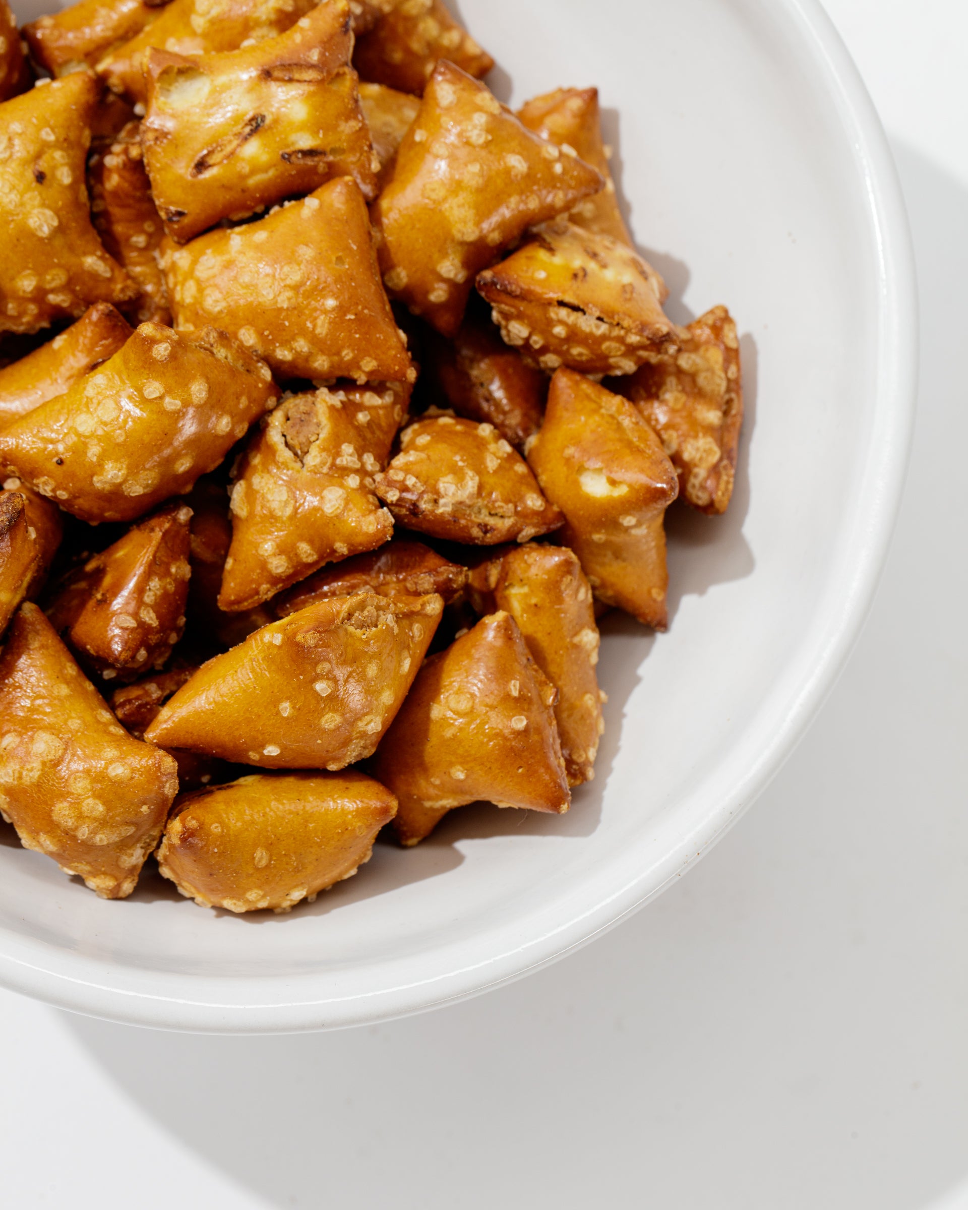 A white Catchall Bowl No. 257 by Montes Doggett, crafted from handmade ceramic dinnerware using traditional techniques, is filled with square-shaped pretzel bites coated in coarse salt. The pretzel bites have a shiny, golden-brown appearance, suggesting they are freshly baked. The bowl is positioned against a light background.