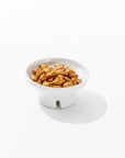 A white Catchall Bowl No. 257 from Montes Doggett rests on a white surface, filled with glossy honey sesame snacks. The bowl, expertly crafted by Peruvian artisans, features a small pedestal base and the snacks are sprinkled with sesame seeds.