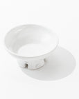 The Catchall Bowl No. 257 from Montes Doggett is a small, white, round ceramic bowl with a glossy finish that stands on three short legs. Artisan crafted from fine quality clays, this handmade ceramic dinnerware piece features a circular emblem with a design in the center. The bowl casts a faint shadow on the white surface beneath it.