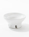 The Catchall Bowl No. 257 by Montes Doggett is a small, white ceramic bowl featuring a wide, shallow shape and two support feet. Artisan-crafted from fine quality clays, the bowl boasts a smooth, glossy finish and features a small logo embossed on one of the feet. The background is plain white.