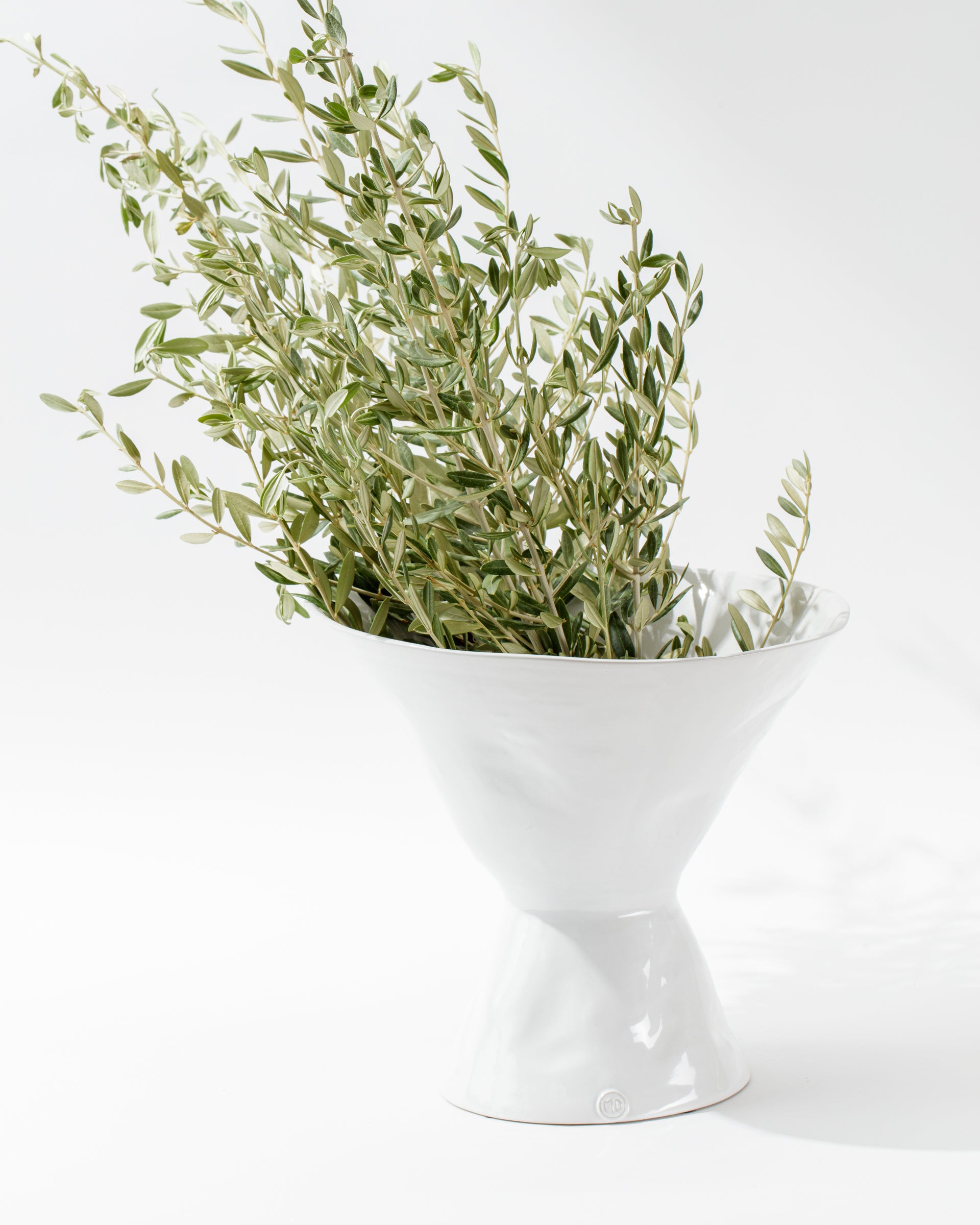 A minimalist, white Catchall Bowl No. 979 with a unique, trumpet-like design holds an arrangement of green leafy branches. Artisan crafted in Peru from high-fired ceramic by Montes Doggett, the bowl stands against a plain white background, emphasizing its clean lines and modern aesthetic.