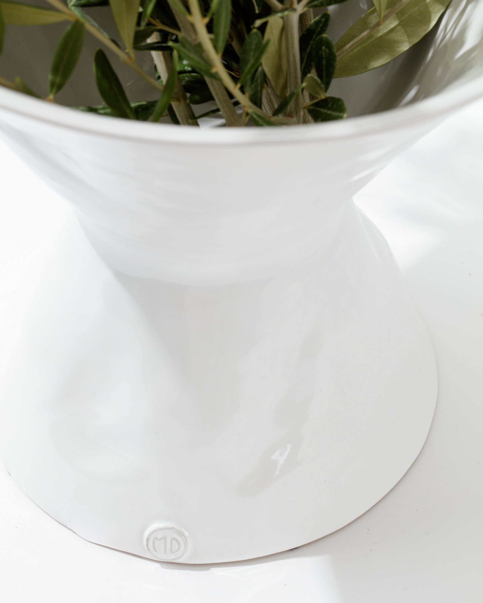 A close-up of a high-fired ceramic Catchall Bowl No. 979 with a wide rim and narrow base from Montes Doggett. Inside the bowl, there are green leafy branches. The glossy surface gleams, set against a bright white background. Artisan crafted in Peru, this piece features a small circular logo with the letters 'MD' at the base.