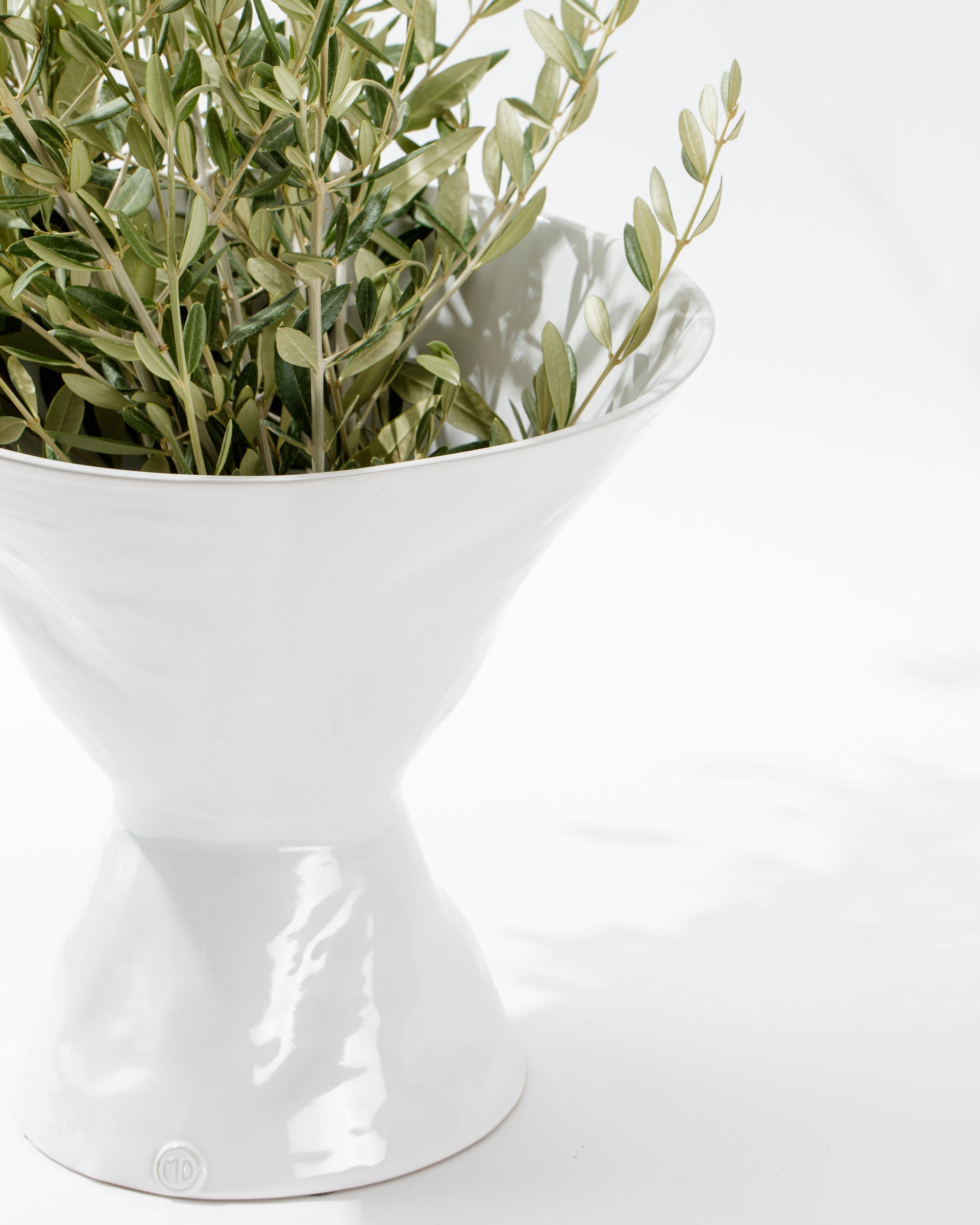 A white ceramic Catchall Bowl No. 979 from Montes Doggett, artisan crafted in Peru, elegantly displays a bundle of green leafy branches. Its high gloss finish reflects light, creating a modern and refined appearance. The plain white background highlights the simplicity and sophistication of the design.