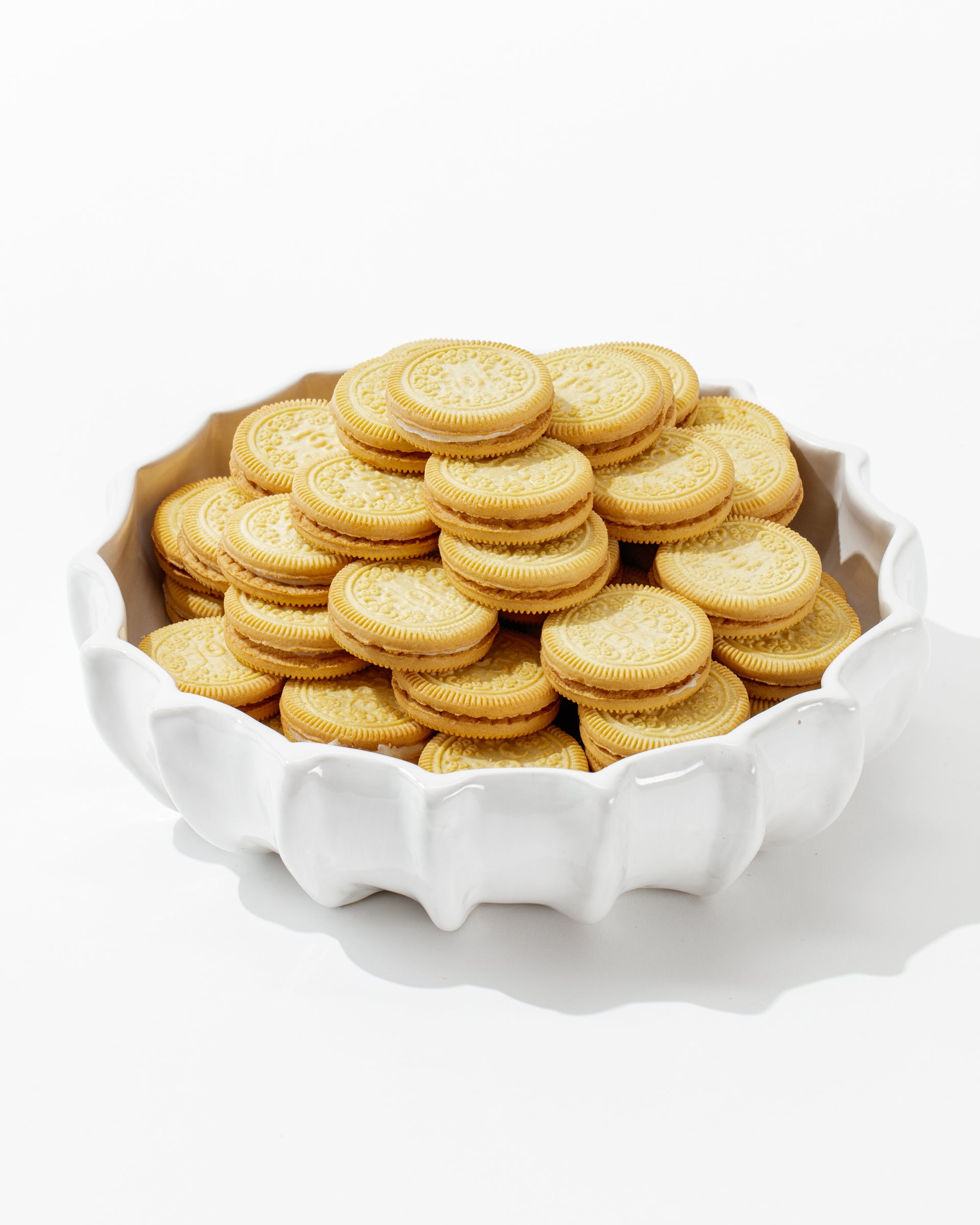 A bowl of golden sandwich cookies, each boasting an intricate pattern on the top biscuit with a layer of cream in the middle, is showcased in Montes Doggett's Bowl No. 727—a white, high-fired ceramic piece featuring a scalloped edge and crafted in Peru. The elegant bowl stands out against a simple white background.