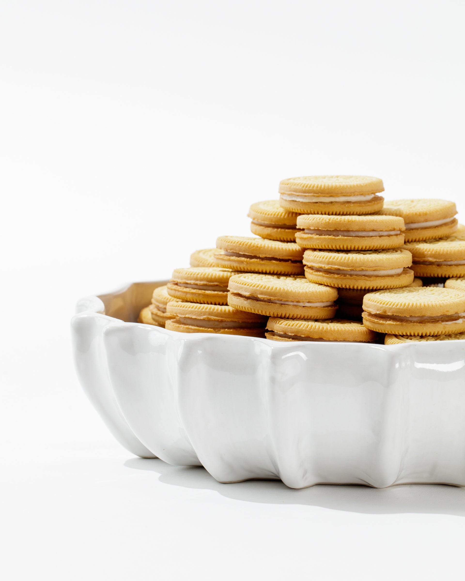 A white, high-fired ceramic dish, known as Bowl No. 727 from Montes Doggett and handmade in Peru, is filled with a stack of golden Oreo cookies. The dish features a wavy, scalloped edge, with the cookies neatly arranged to showcase the creamy filling visible between the two biscuits. The background is plain white.