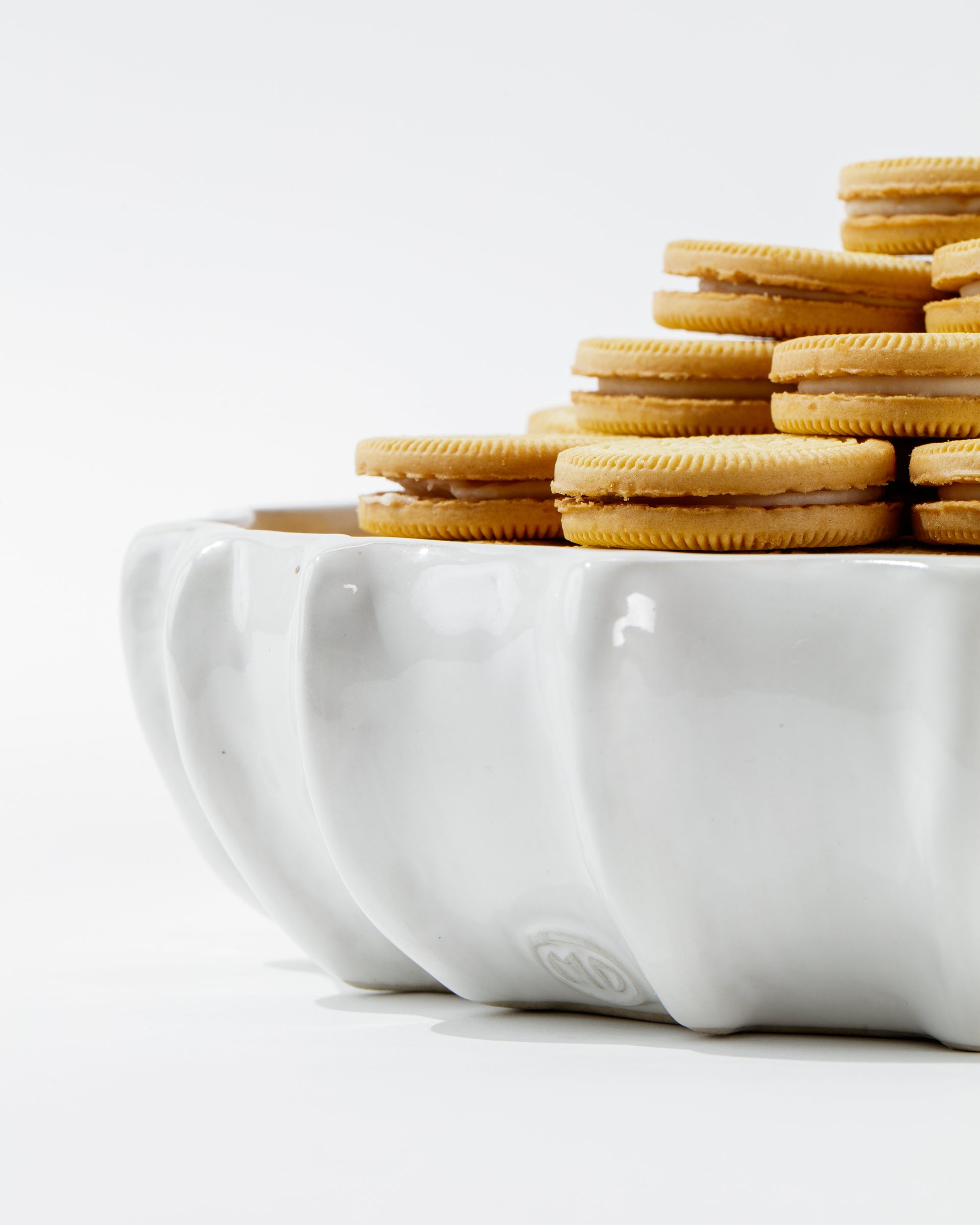 Bowl No. 727 by Montes Doggett, a white, high-fired ceramic piece handcrafted in Peru, is elegantly filled with a stack of cream-filled sandwich cookies arranged neatly. The bowl features a wavy, scalloped edge design that stands out against the plain white background, drawing attention to both the handmade ceramic bowl and the delicious cookies.